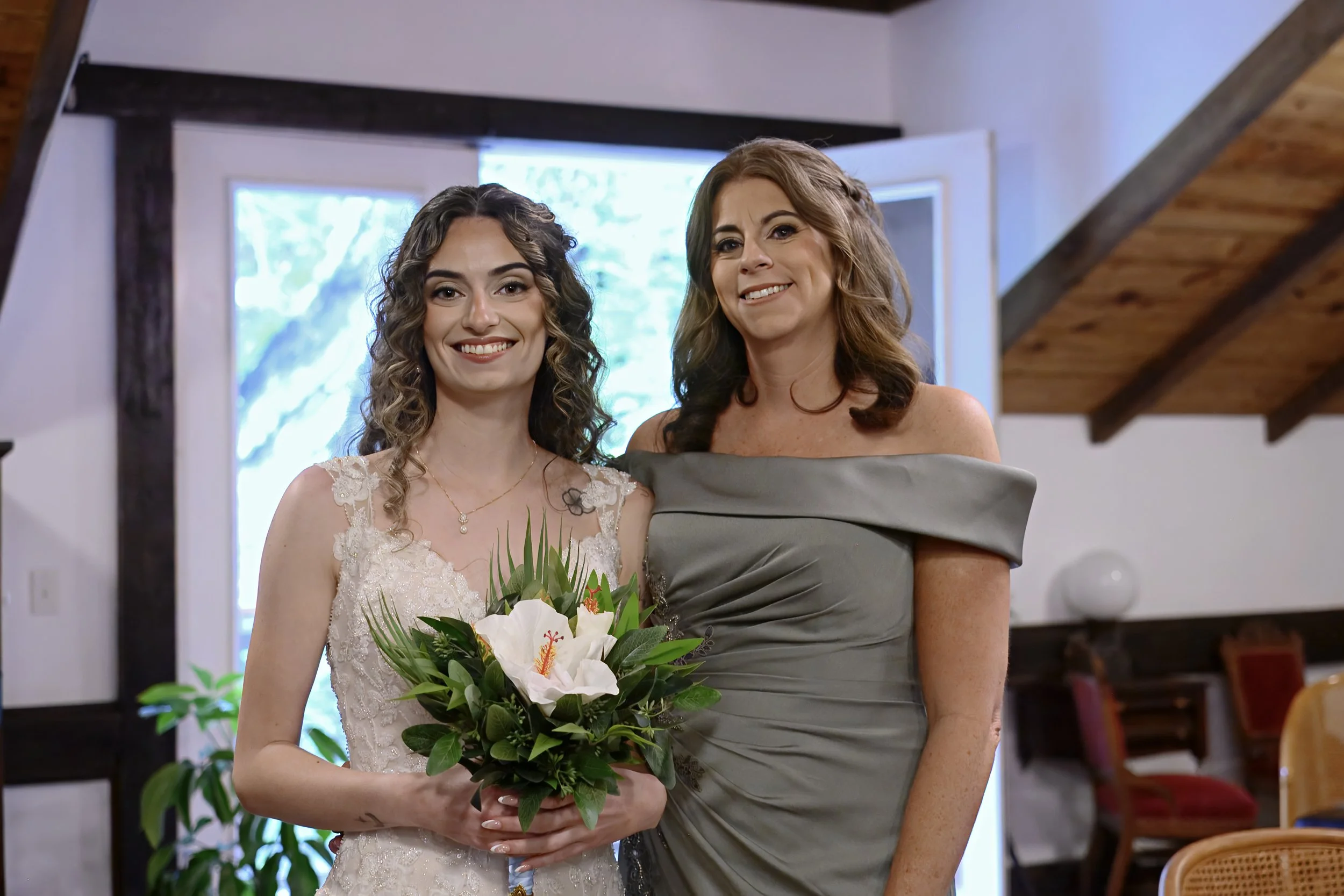 Two women, one in a wedding dress holding a bouquet of flowers and the other in a gray dress, are standing close together indoors, smiling at the camera.