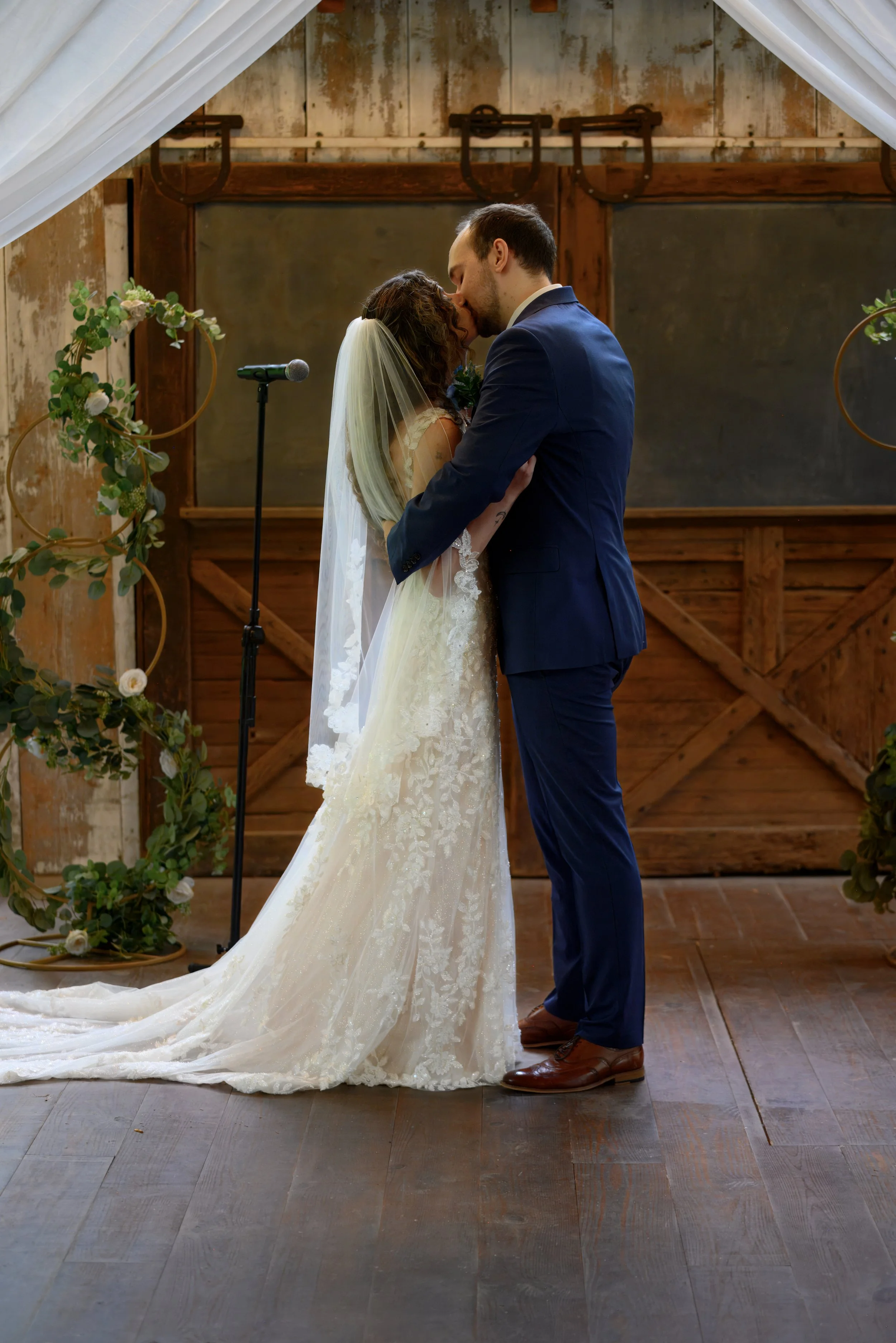 A bride and groom share a kiss during their wedding ceremony inside a rustic venue. The bride wears a lace wedding gown and veil, while the groom is dressed in a dark blue suit. Floral decorations adorn the arch behind them, with a wooden door and a 