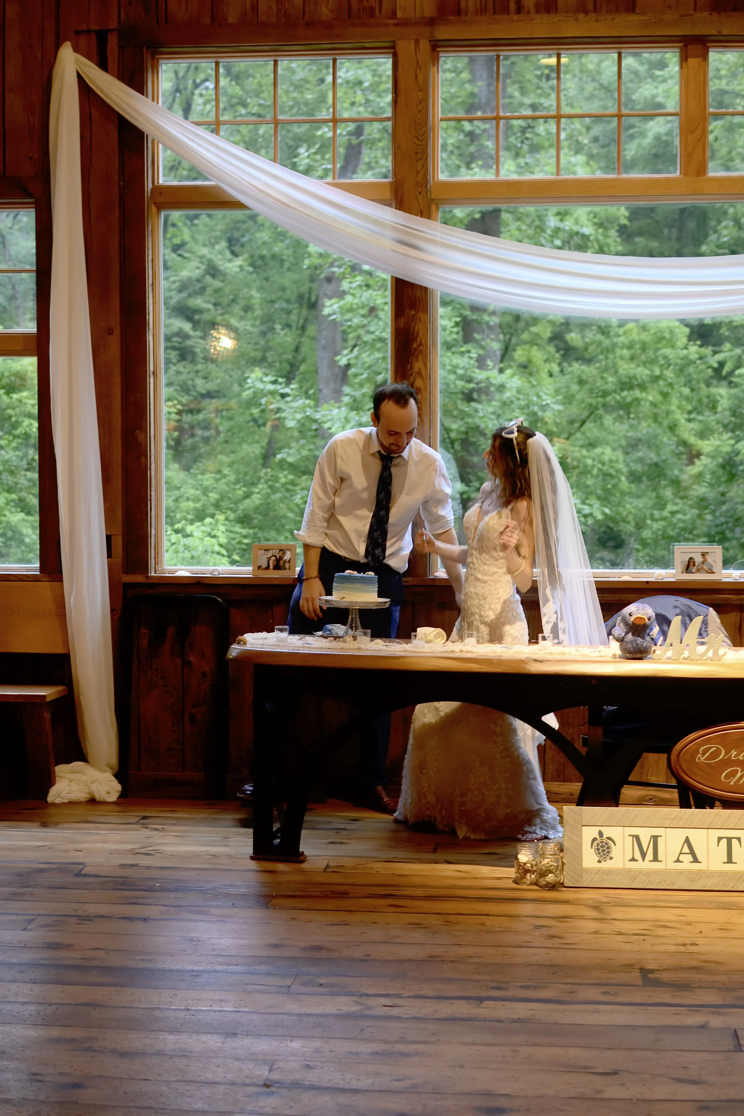A bride and groom at their wedding reception standing behind a table with a cake. The groom is leaning over the table, and the bride is looking at him. The background features large windows showing green trees outside, with wooden walls and flooring 