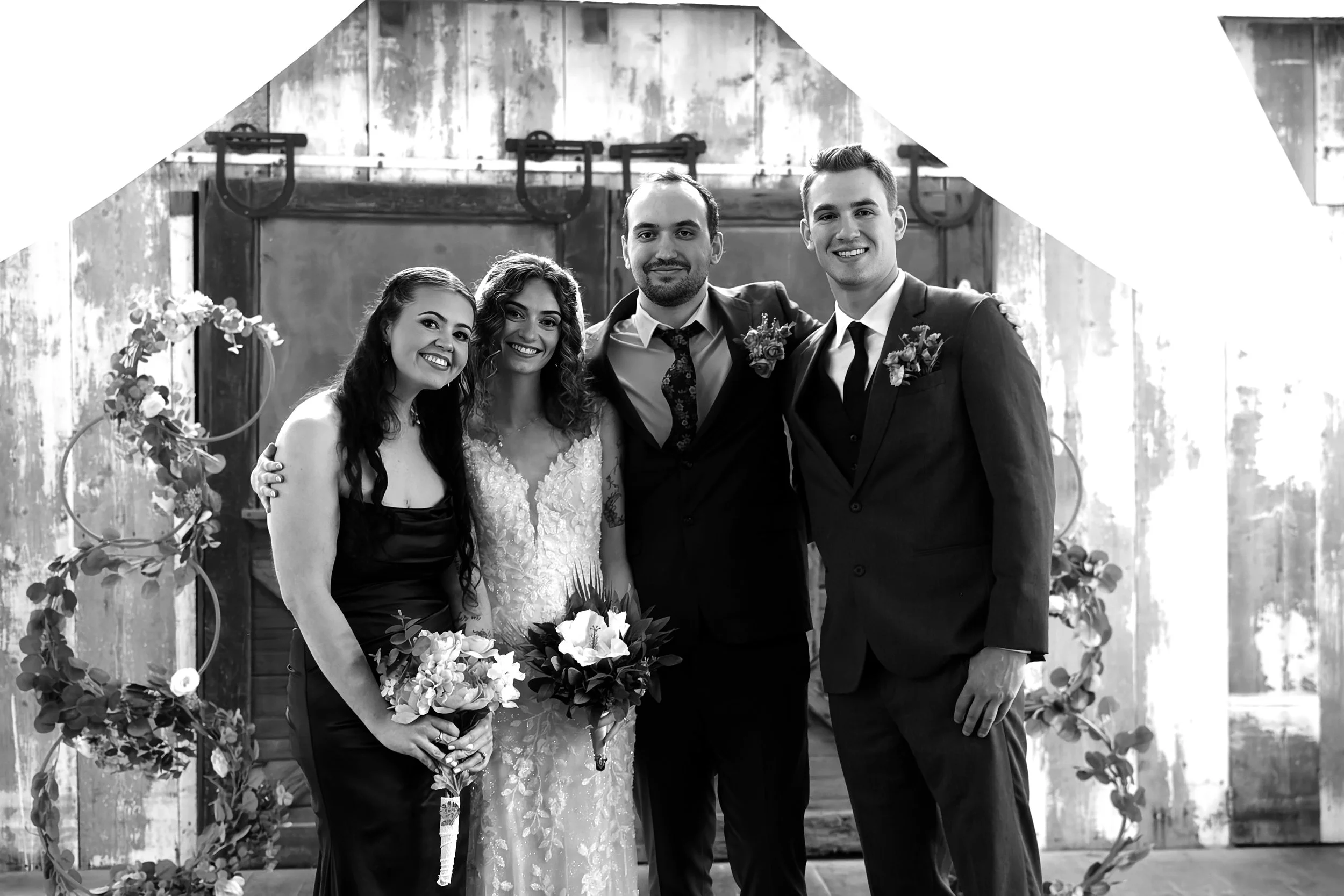 Black and white photo of a wedding party with four people standing in front of a rustic background with wooden panels and a sliding barn door. The woman in the center is wearing a wedding dress, holding a bouquet, and standing next to the man in a su