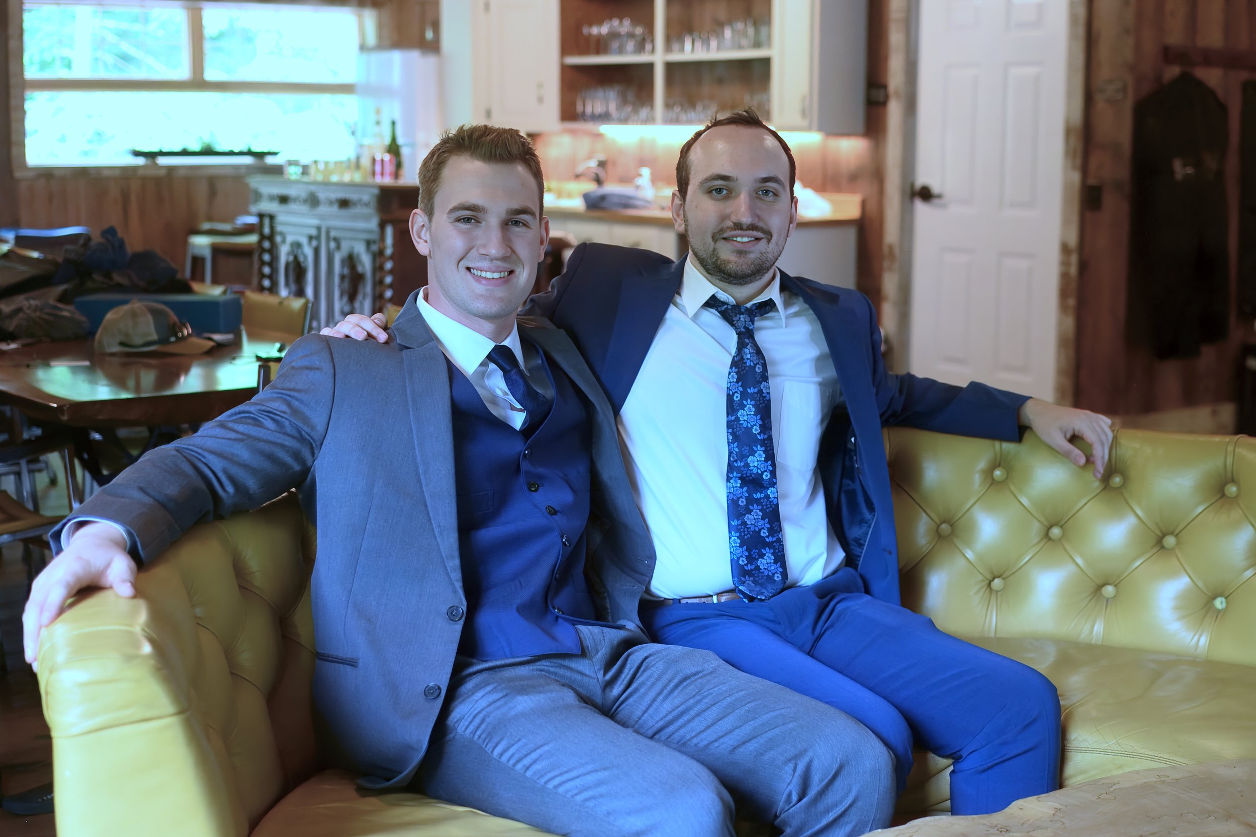 Two men in suits sitting close together on a yellow leather couch, smiling at the camera in a cozy, rustic-style room with wood paneling, a window, and a kitchen area in the background.
