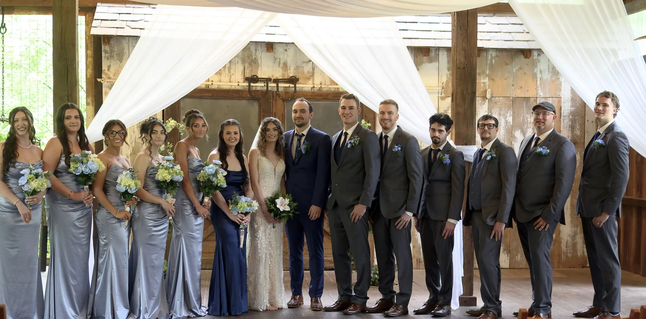 A wedding party with bridesmaids in matching blue dresses and groomsmen in suits, standing together indoors with rustic wooden walls and white drapes draped overhead.