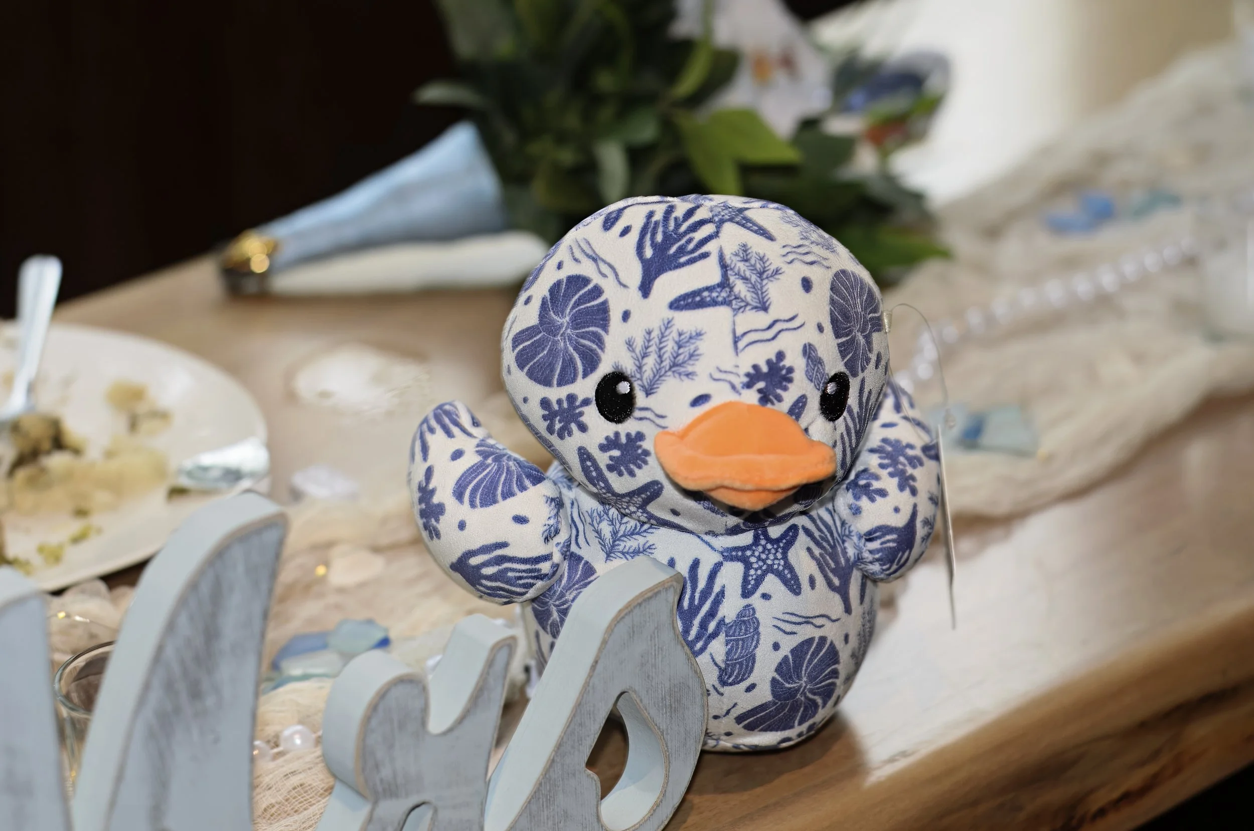 A plush duck toy with a blue floral pattern on white fabric, sitting on a wooden table near a white decorative item.