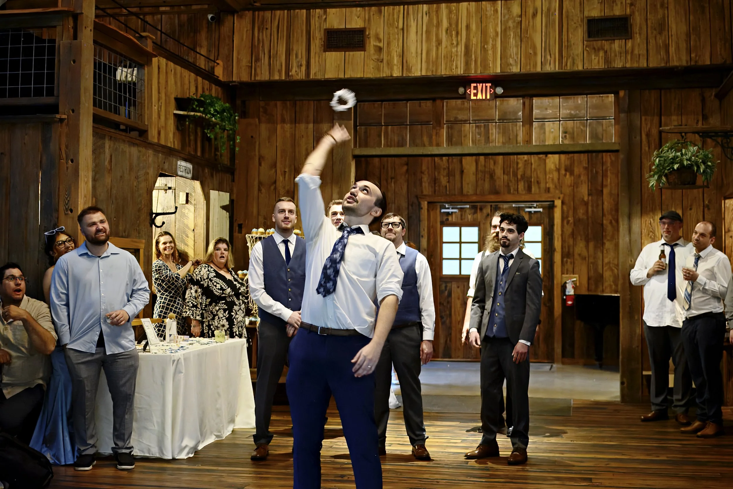A group of people at a social gathering or celebration in a rustic wooden hall. One man is tossing a napkin into the air, and others are watching and smiling. The setting includes wooden walls, indoor plants, and an EXIT sign.