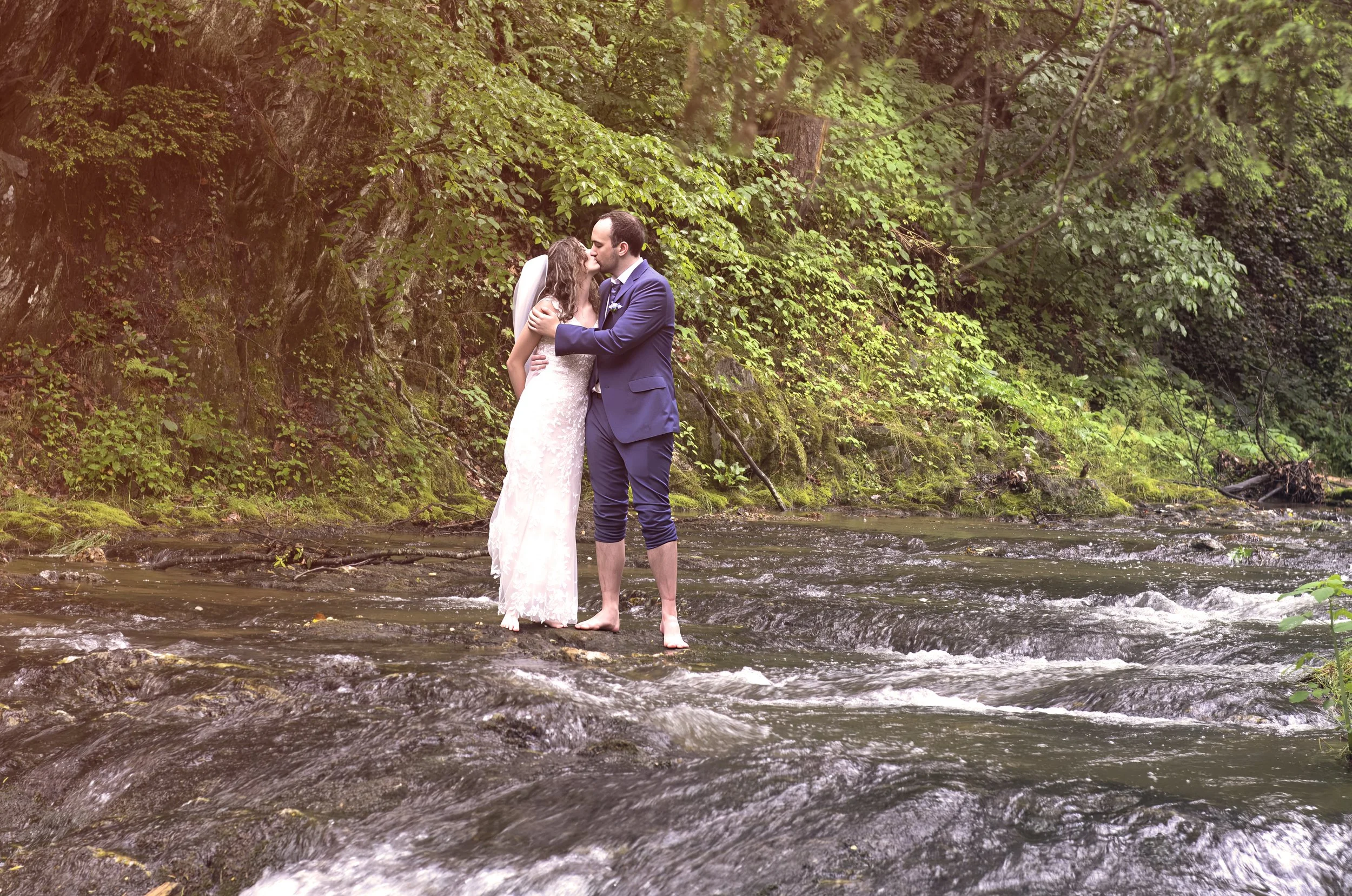 A bride and groom kissing in a shallow river surrounded by lush green trees and moss-covered rocks.