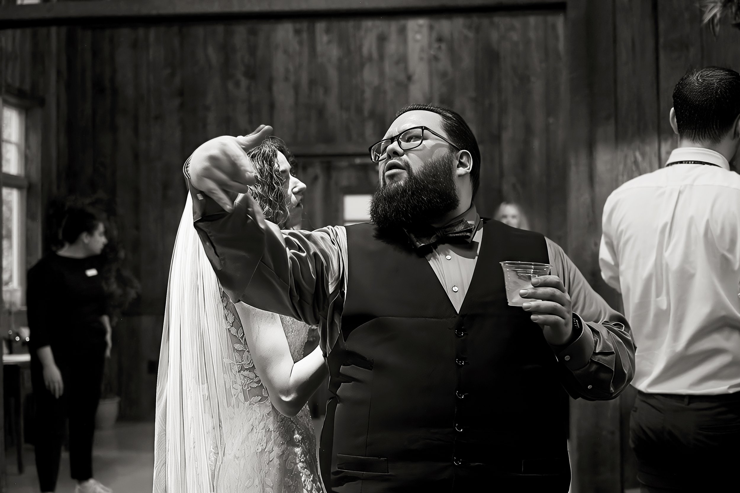 Black and white photo of a man with glasses and a beard wearing a tuxedo, dancing with a woman in a wedding dress, at a wedding reception. Other people are visible in the background.