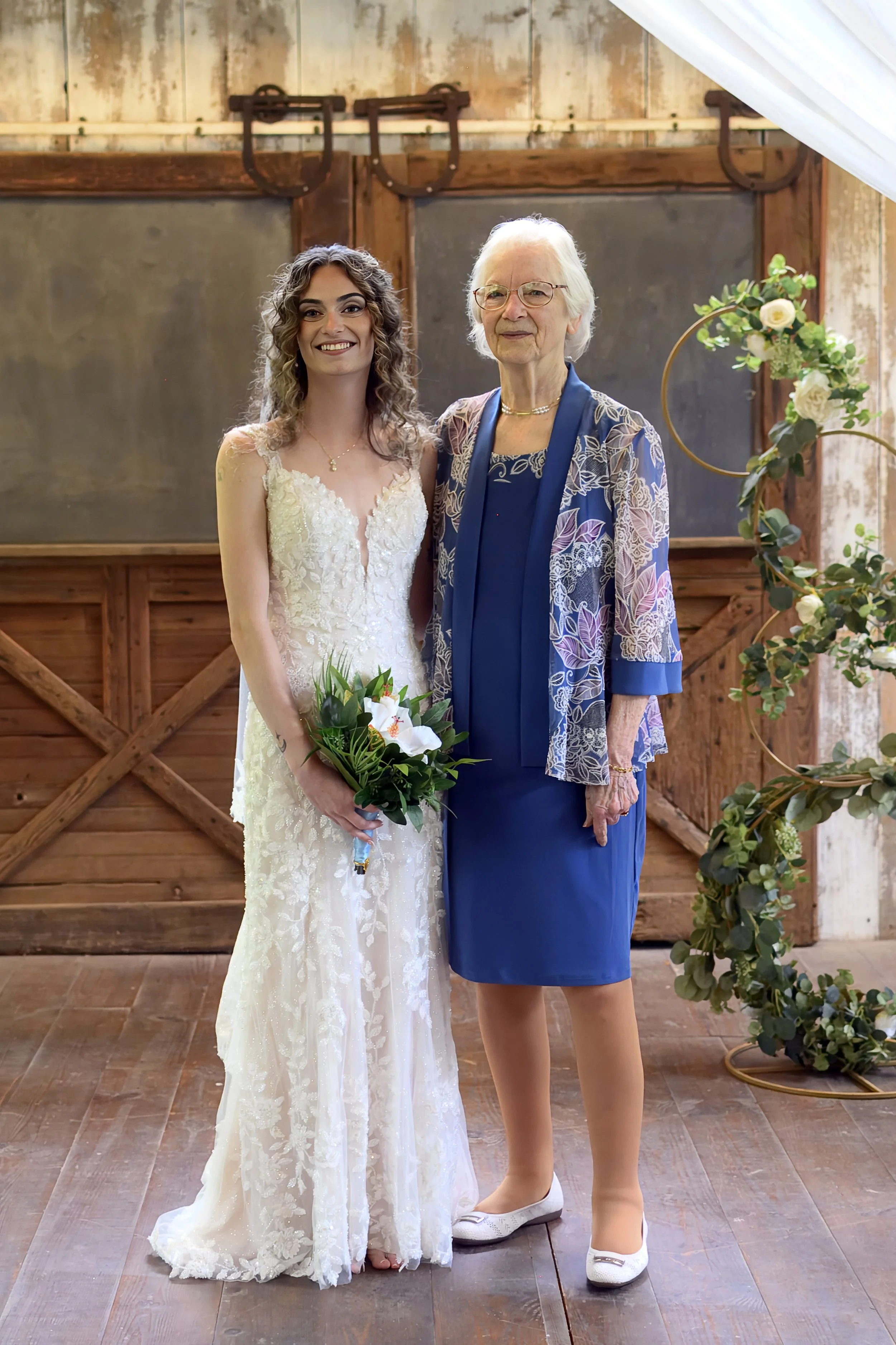 A bride in a white lace wedding dress holding a bouquet standing next to an older woman in a blue dress and patterned blazer, inside a rustic wooden barn decorated with greenery and flowers.