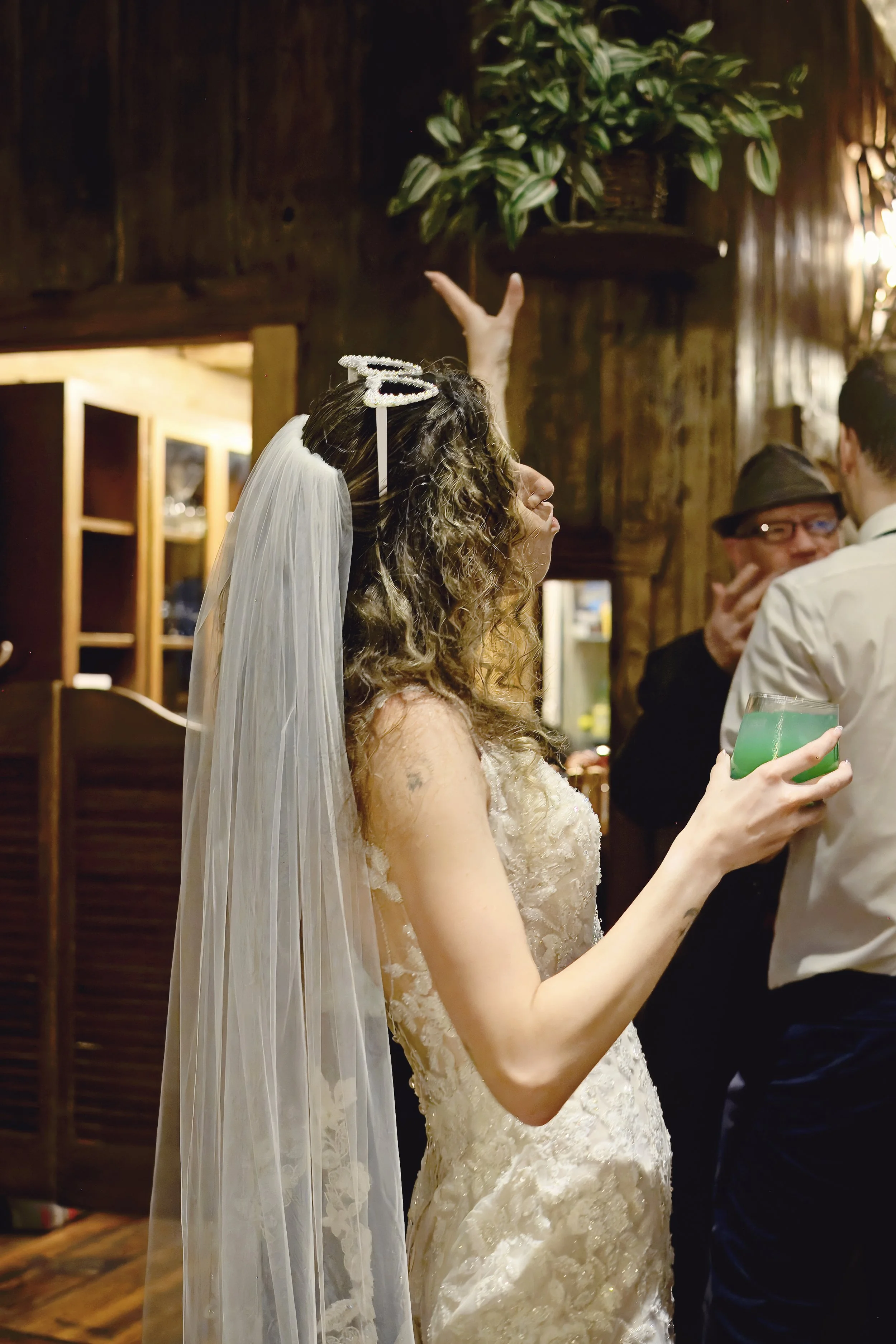 A bride with long curly hair wearing a lace wedding dress and a veil, holding a green drink in her hand, at a wedding reception with wooden walls and other guests in the background.