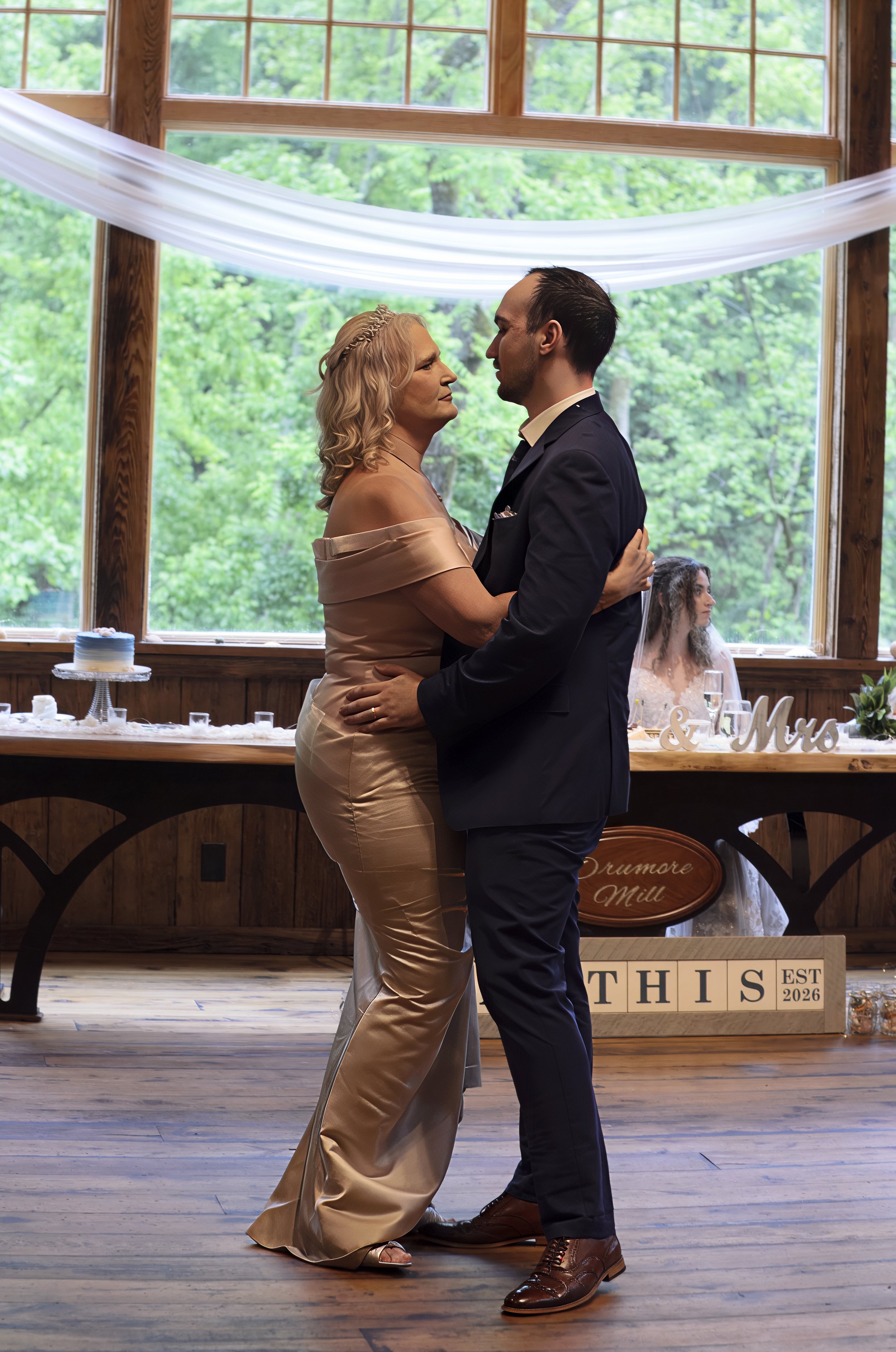 A woman and a man are dancing together at a wedding reception, with trees visible through large windows in the background. The woman is wearing a satin, off-shoulder gown, and the man is wearing a dark suit. A bride in a wedding dress is sitting at a