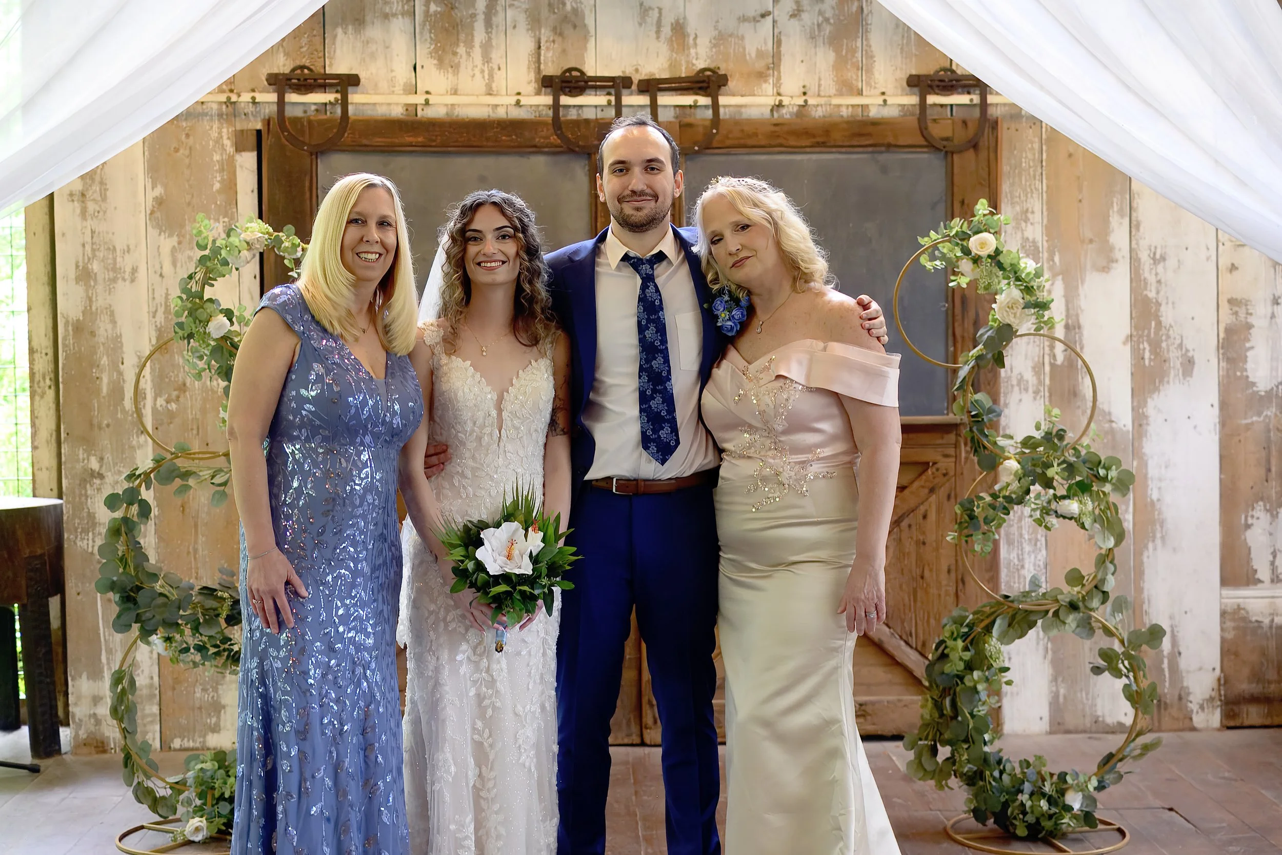 A group of five people at a wedding, including a bride in a white gown holding a bouquet, a groom in a navy suit, two women in formal dresses, and a woman in a pastel yellow dress, standing in front of a rustic wooden backdrop with greenery and white