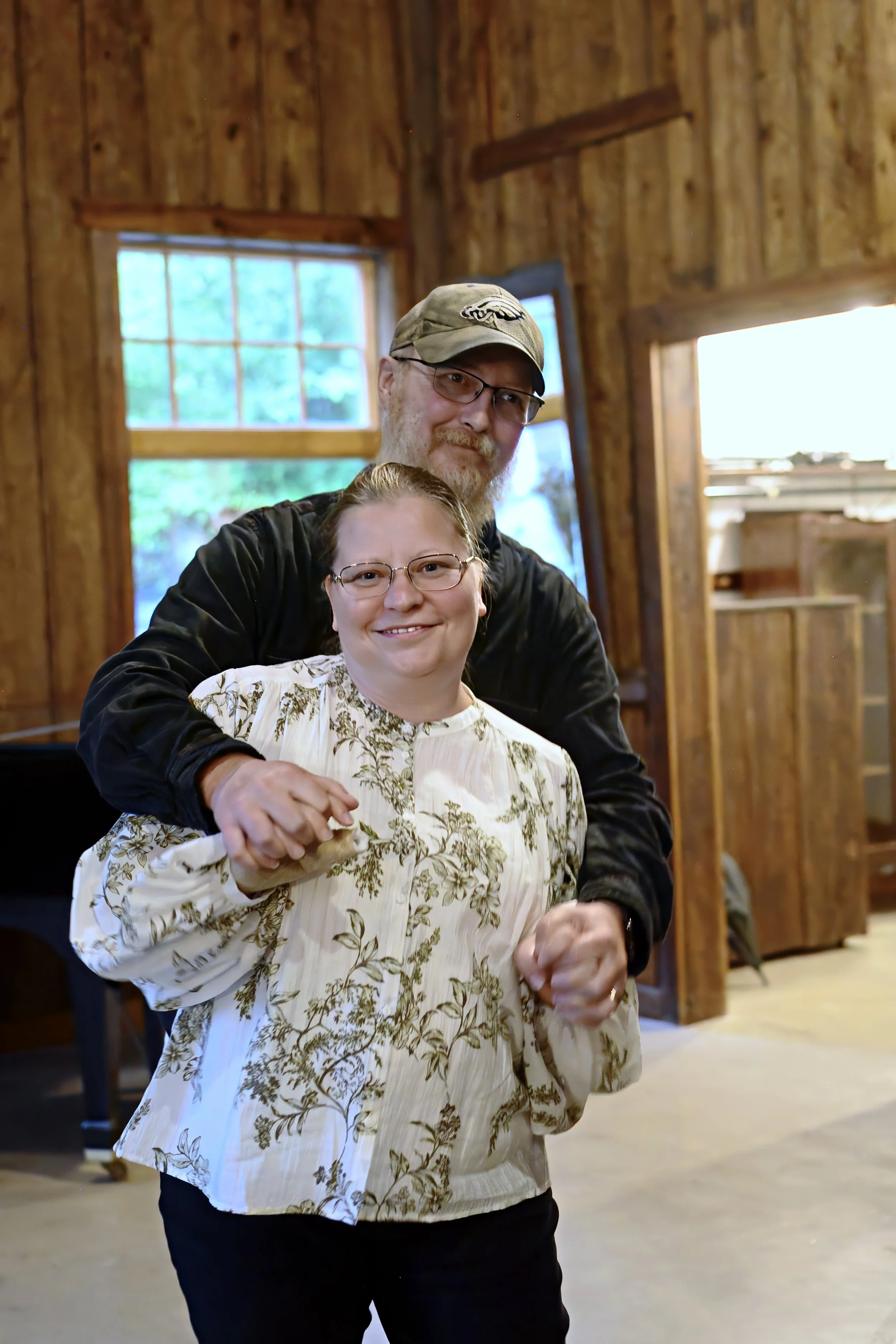 A man and woman smiling and holding hands indoors with wooden walls and windows in the background.