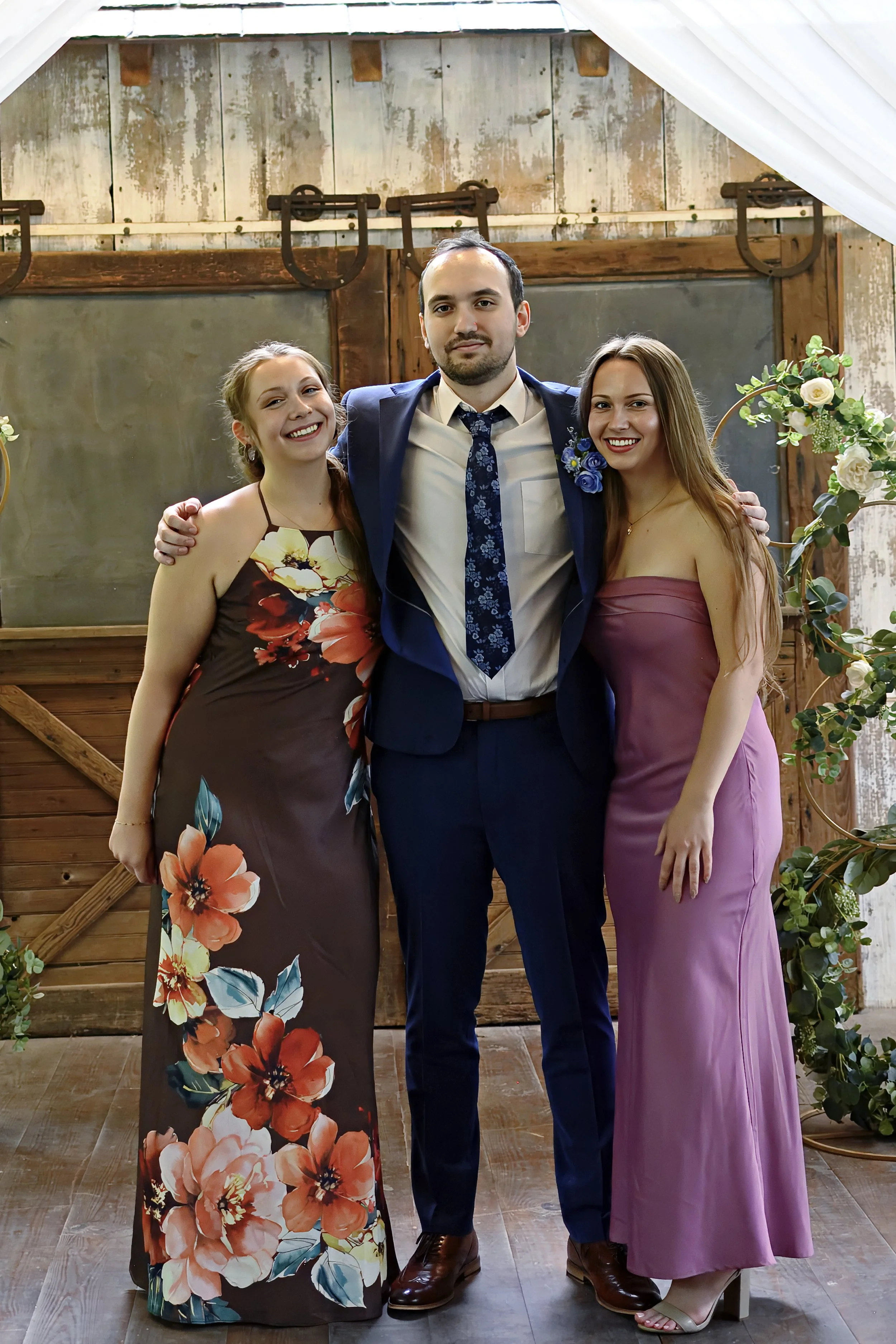 Three people, two women and one man, standing close together and smiling at a rustic wooden backdrop decorated with greenery and flowers, possibly at a wedding or celebration.
