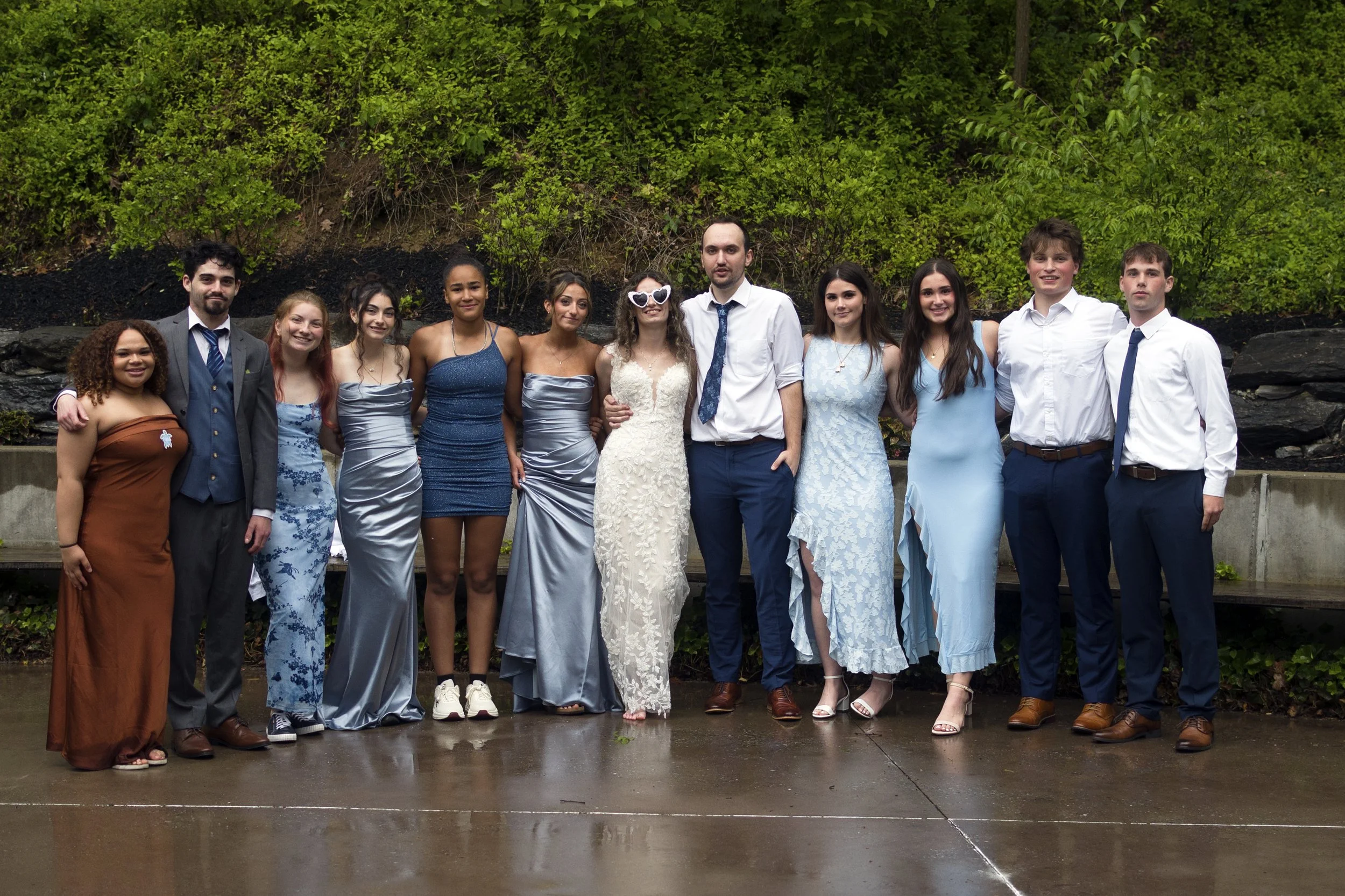A group of eleven young adults in formal attire standing outdoors on a concrete surface, with a background of green trees and bushes.