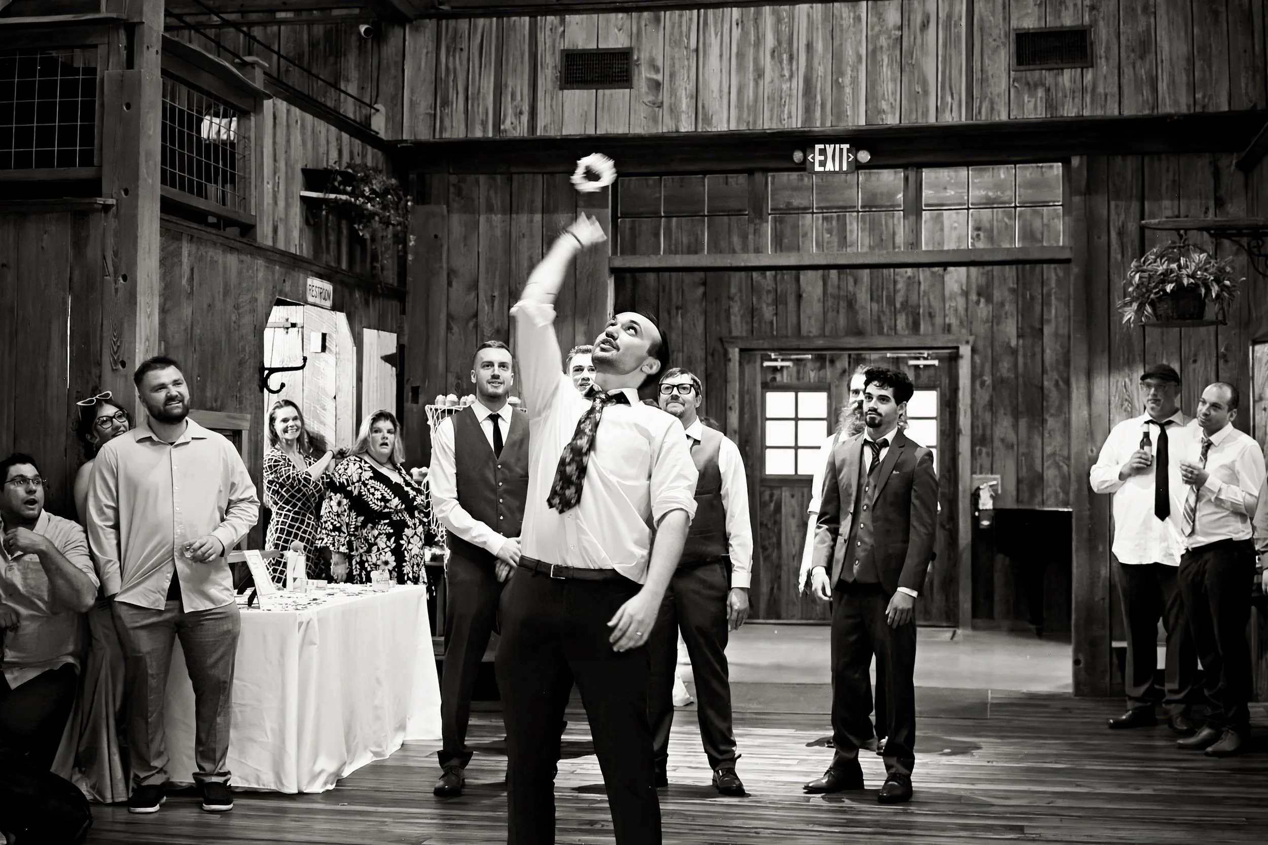 A group of people at a wedding reception inside a rustic wooden building, with a man in a white shirt and tie in the center, throwing a bouquet in the air, surrounded by guests watching and smiling.