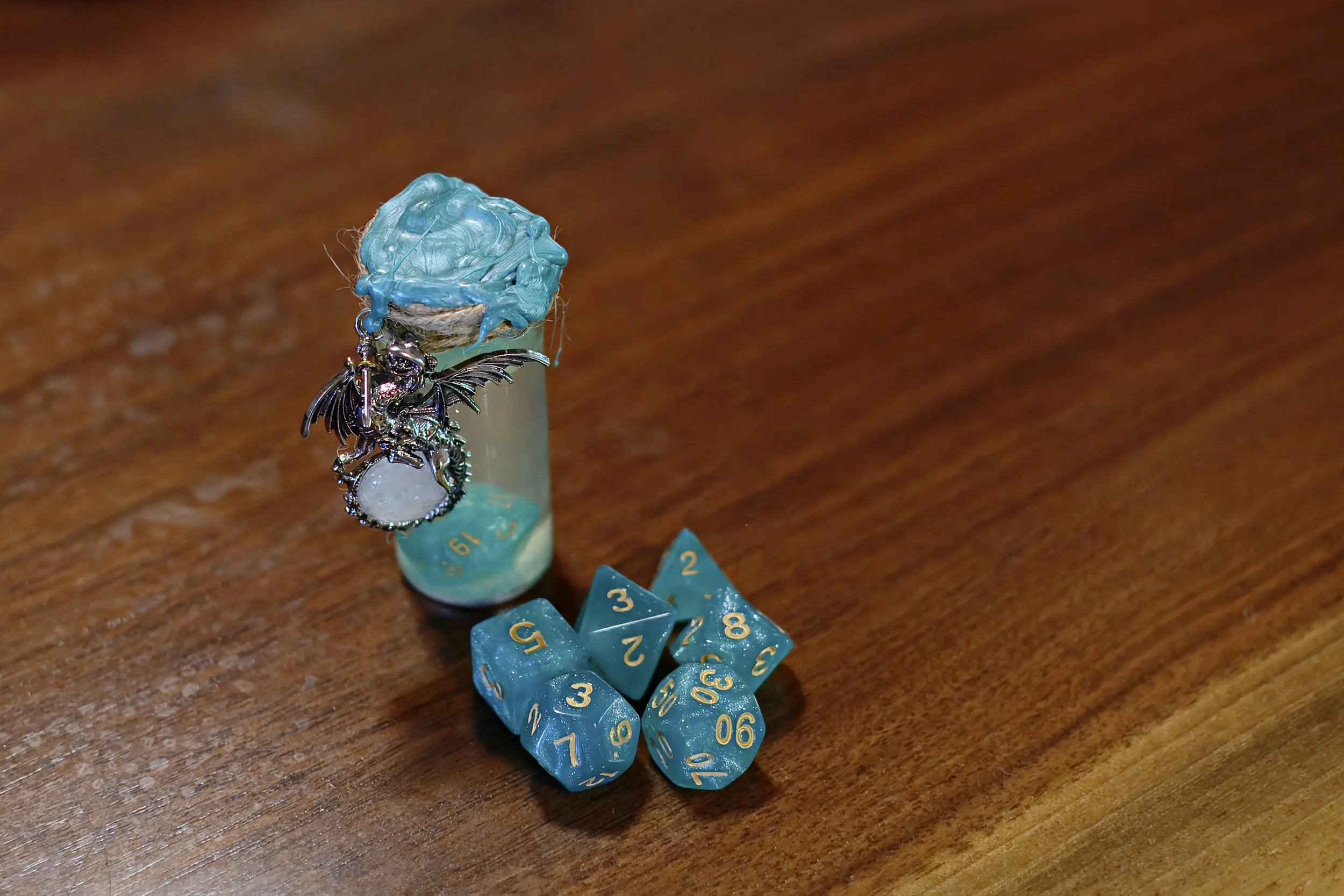 A tabletop gaming setup featuring a turquoise D20 die, a set of five turquoise polyhedral dice, a small clear plastic vial with a blue lid, a silver dragonfly charm, and a blue candy or ornament wrapped in blue paper topped with blue paint.