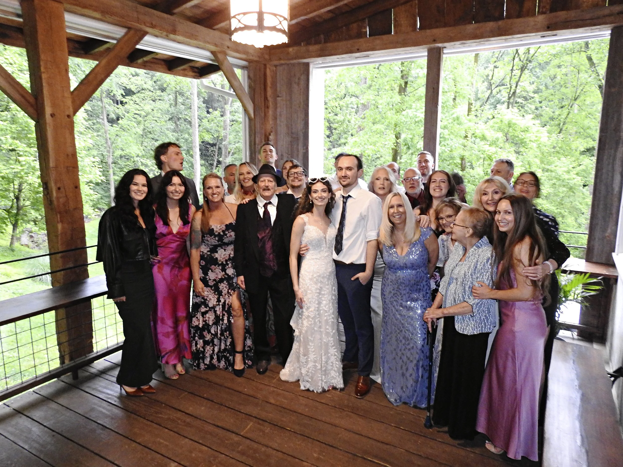 A group of people at a wedding, with the bride and groom in the center, inside a wooden structure surrounded by green trees.