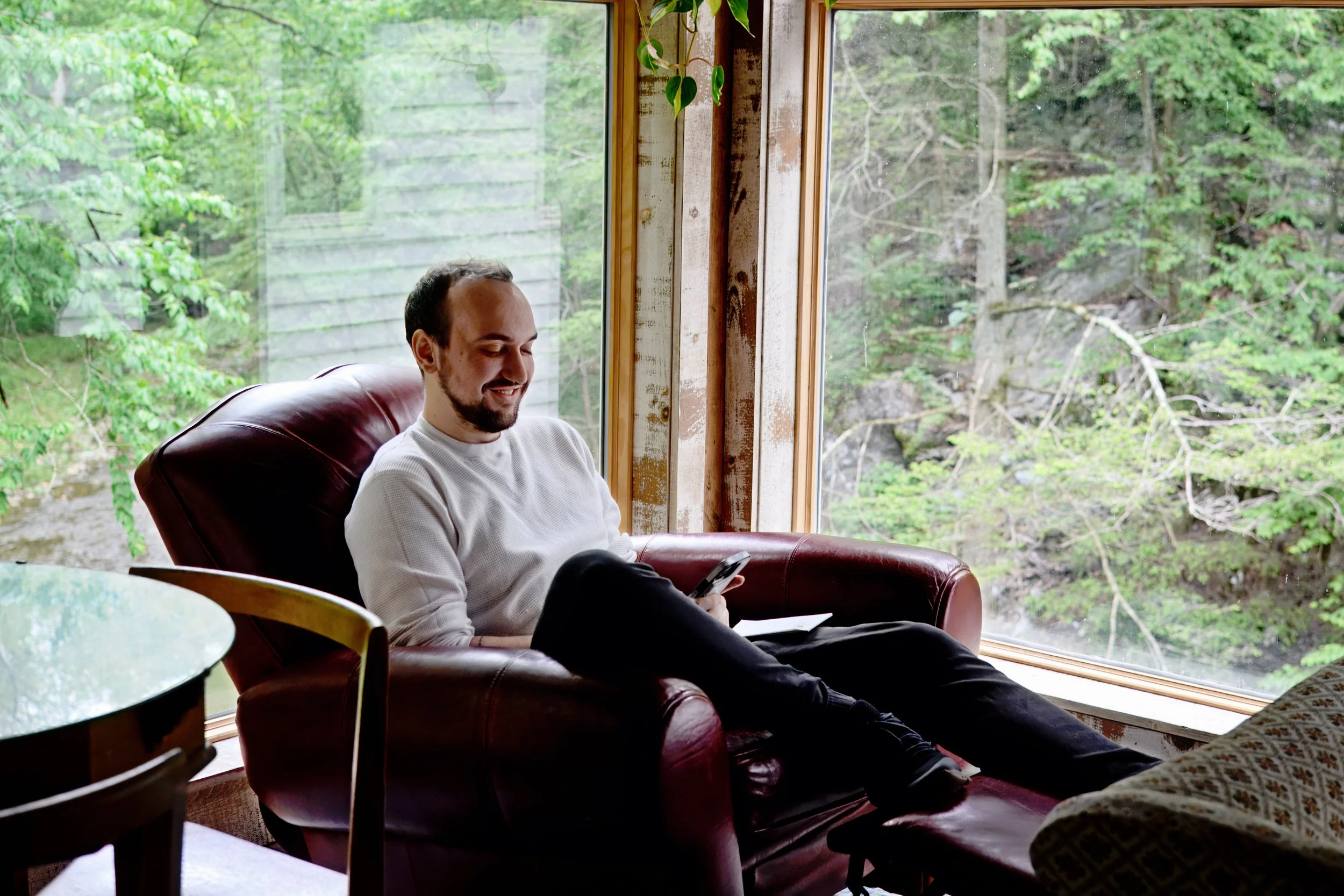A man with a beard sitting on a red leather armchair, smiling and looking at his phone, near a large window with a view of green trees outside.