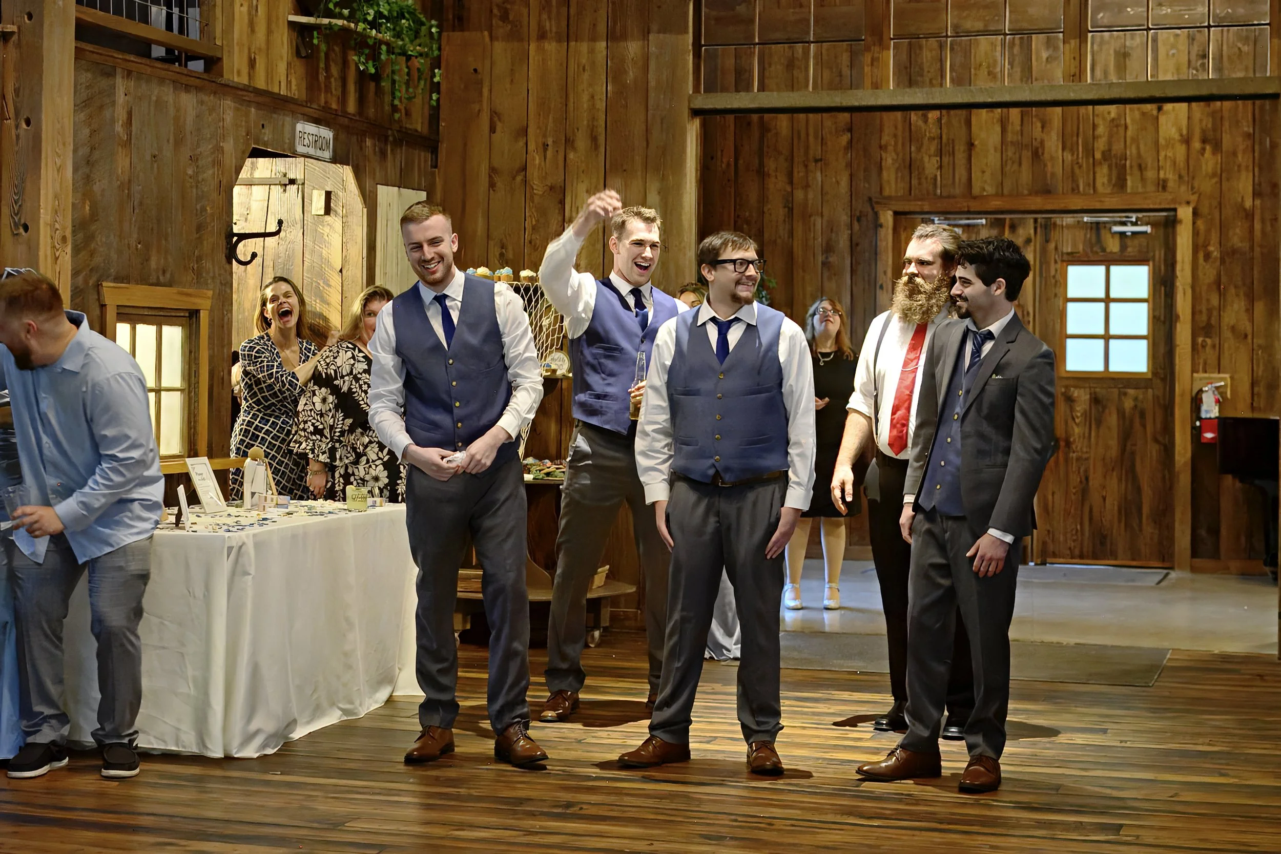 A group of people dressed in formal attire enjoying a celebration inside a rustic wooden venue. Some are smiling and laughing, others are engaged in conversation. There is a table with a display of small items or gifts on the left side of the image.
