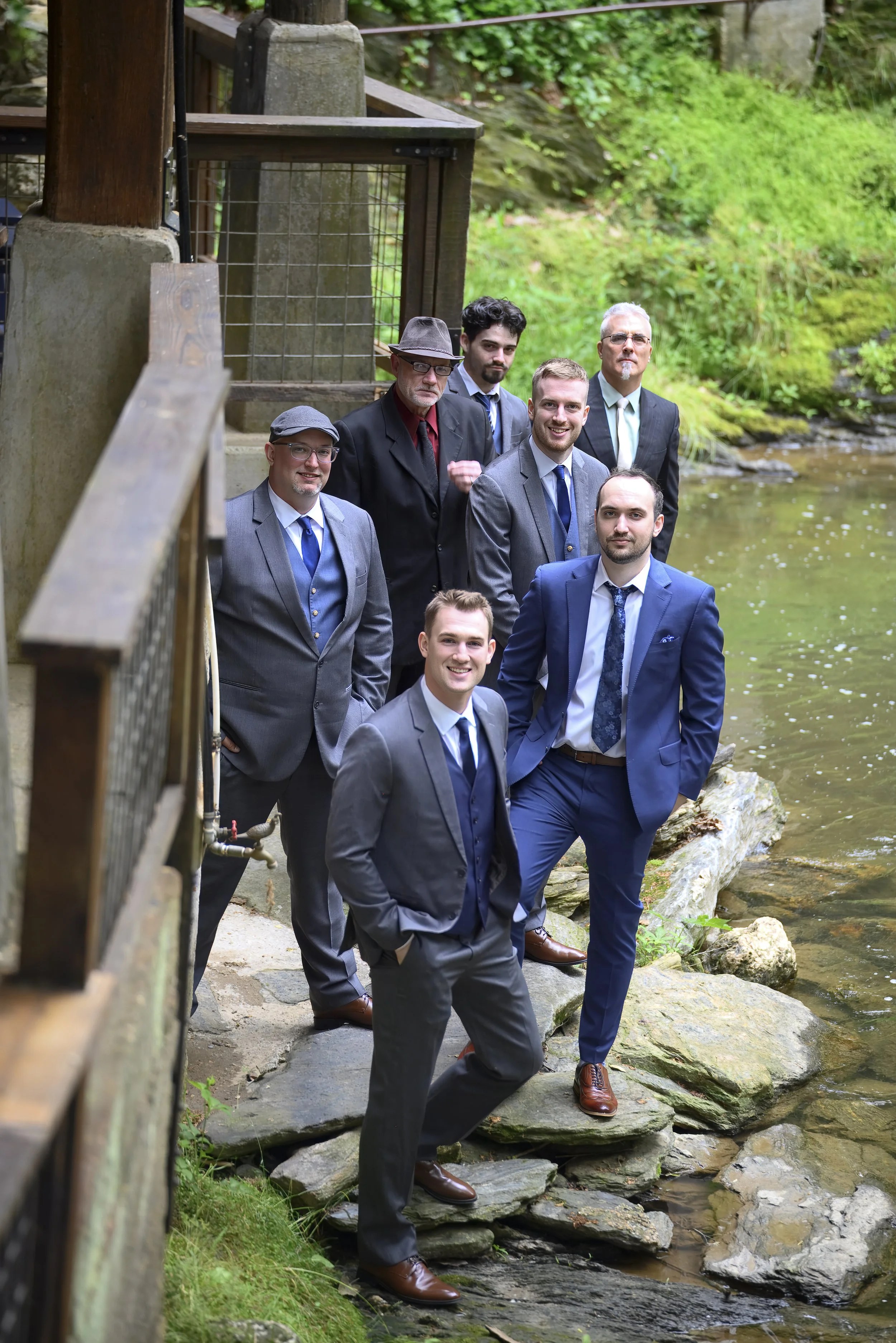 Group of seven men in formal suits standing on rocks by a creek in a lush green forest, with a wooden bridge railing visible on the left side of the image.