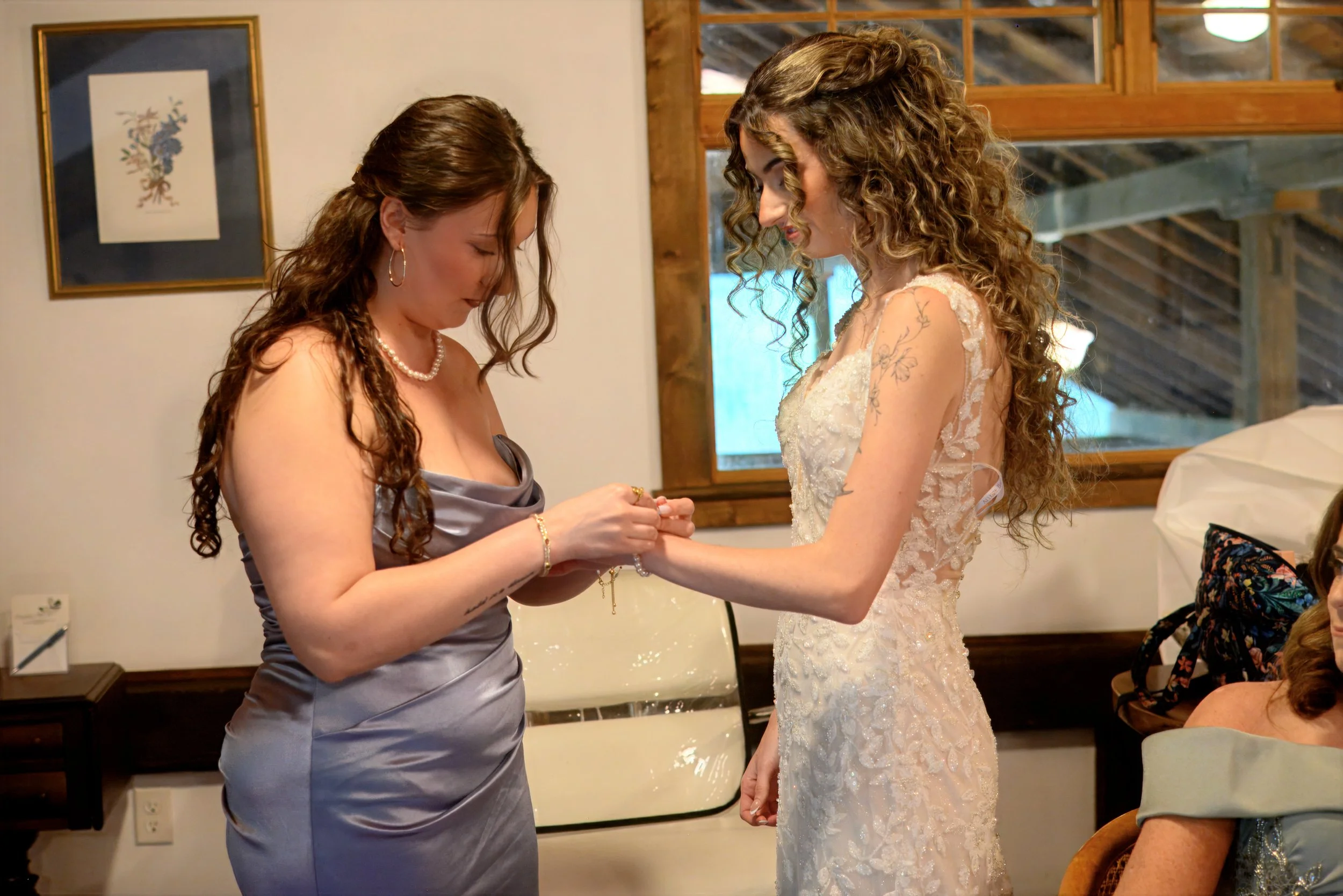 Two women exchanging a bracelet, one in a gray dress and the other in a wedding gown, inside a room with a window and framed artwork on the wall.