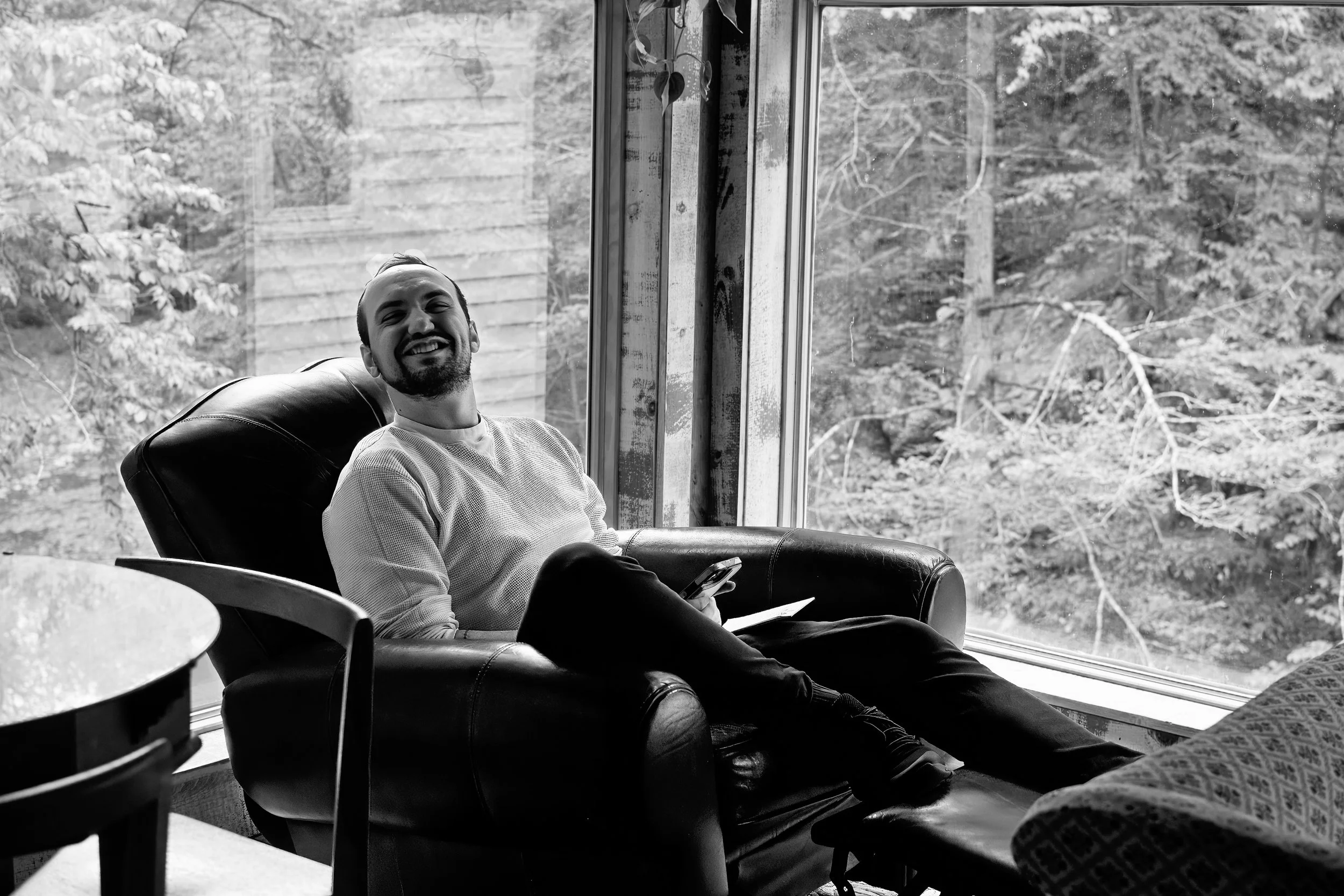 A smiling man sitting in a chair inside a house, looking out a window at trees and a wooden fence outside. He is holding a smartphone in his lap.
