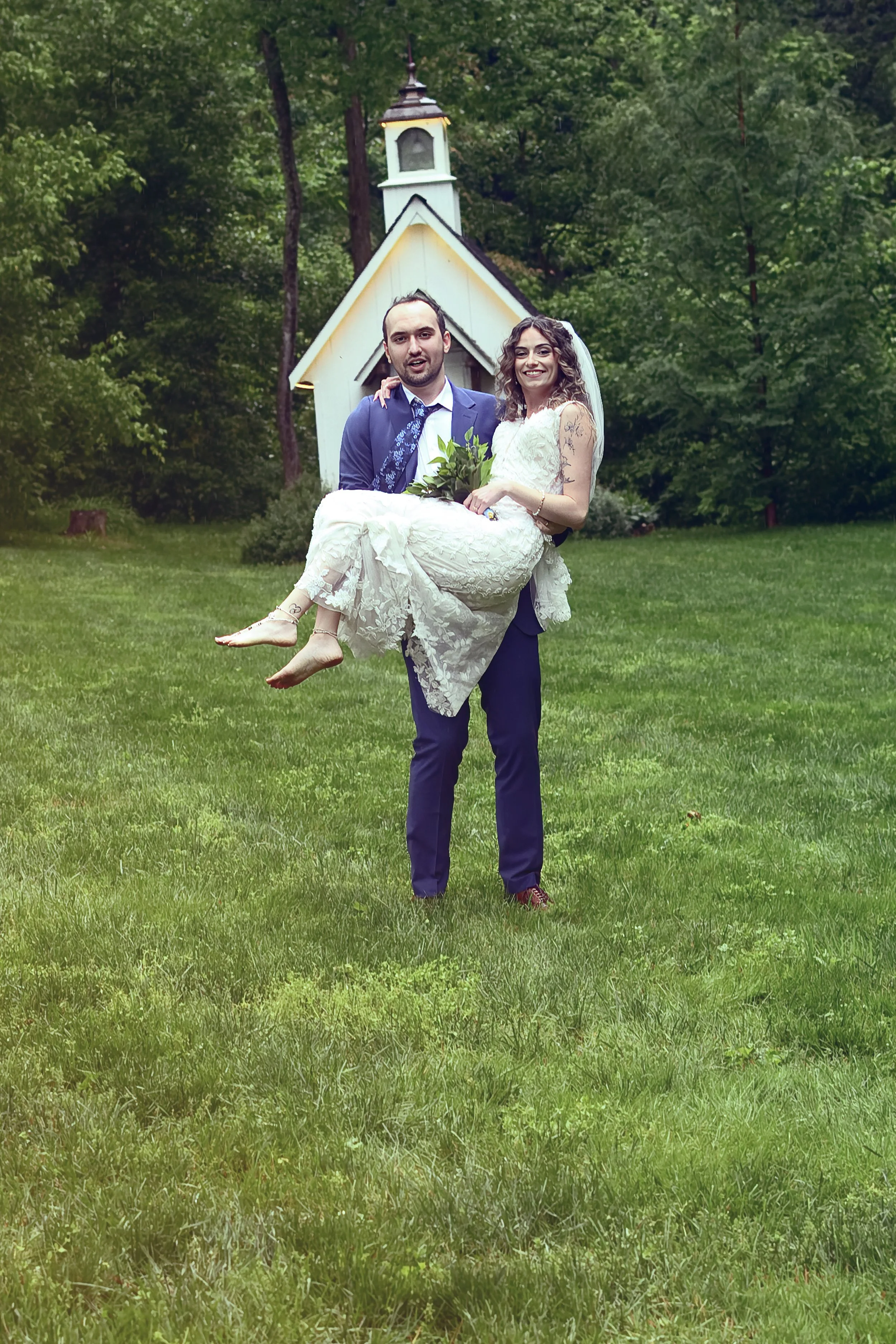 A groom carrying a bride in a white wedding dress with tattoos on her arm, standing on green grass in front of a small white chapel surrounded by trees.