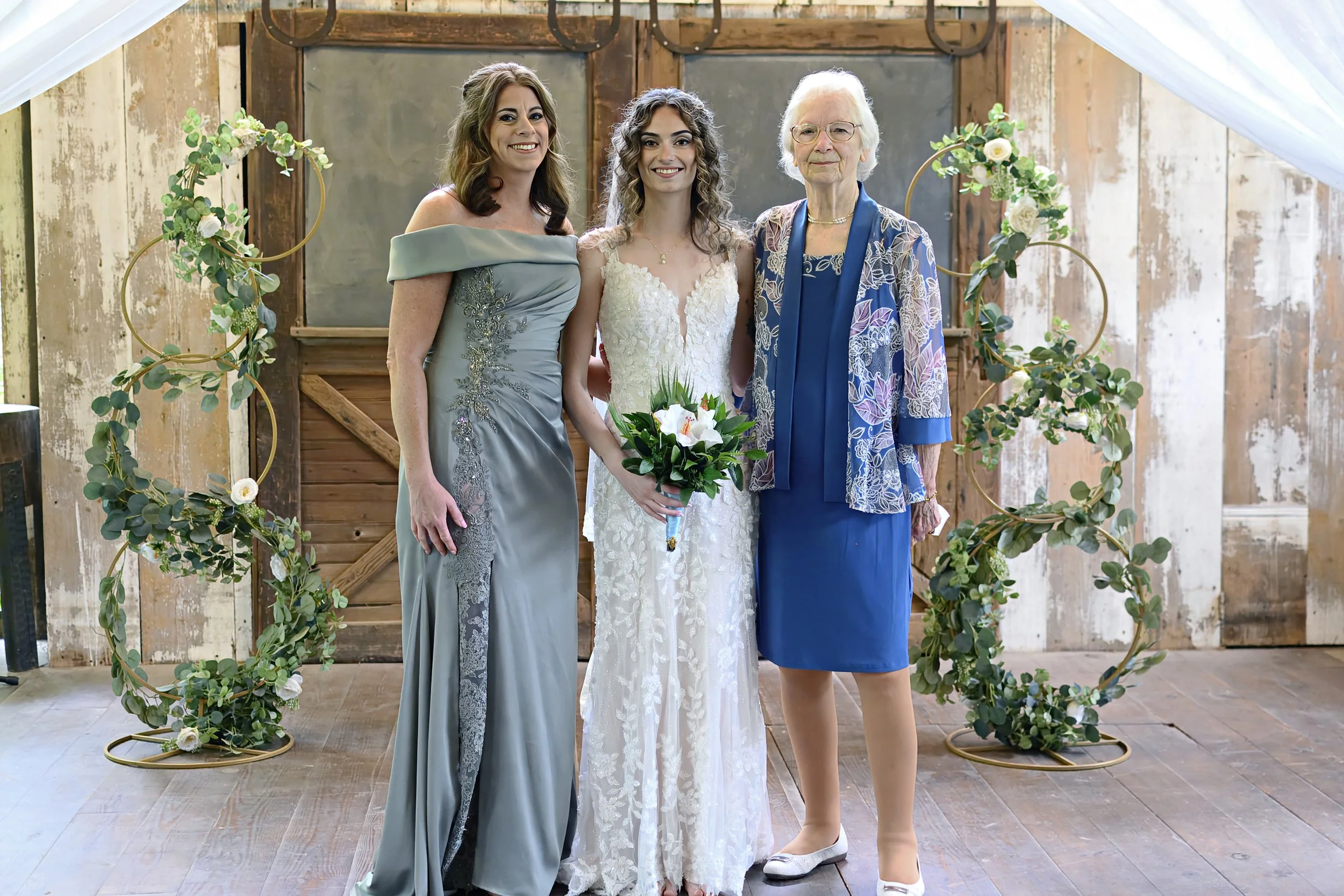 Three women, one in a wedding dress holding a bouquet, standing under a floral arch, smiling at a wedding ceremony. The woman on the left wears a gray dress, the woman on the right wears a blue dress, and an older woman on the far right wears a blue 