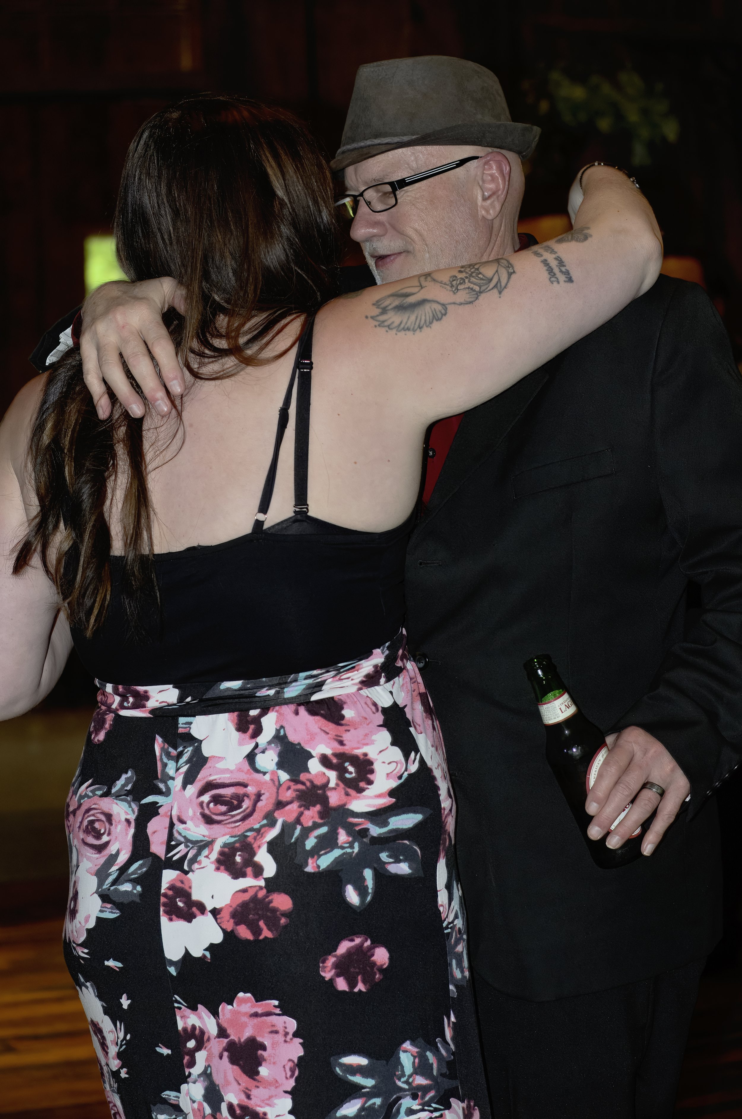 A couple dancing closely at a social event, with the woman wearing a floral skirt and the man holding a beer bottle.