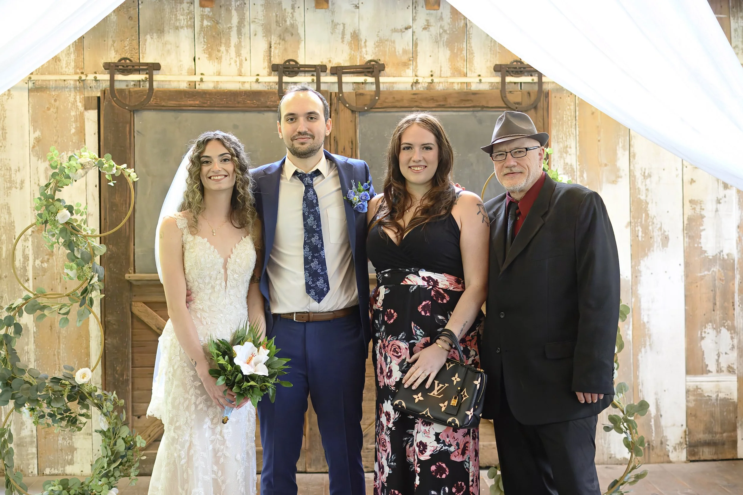 A group of four people at a wedding, standing in front of a rustic wooden backdrop with greenery and floral decorations.