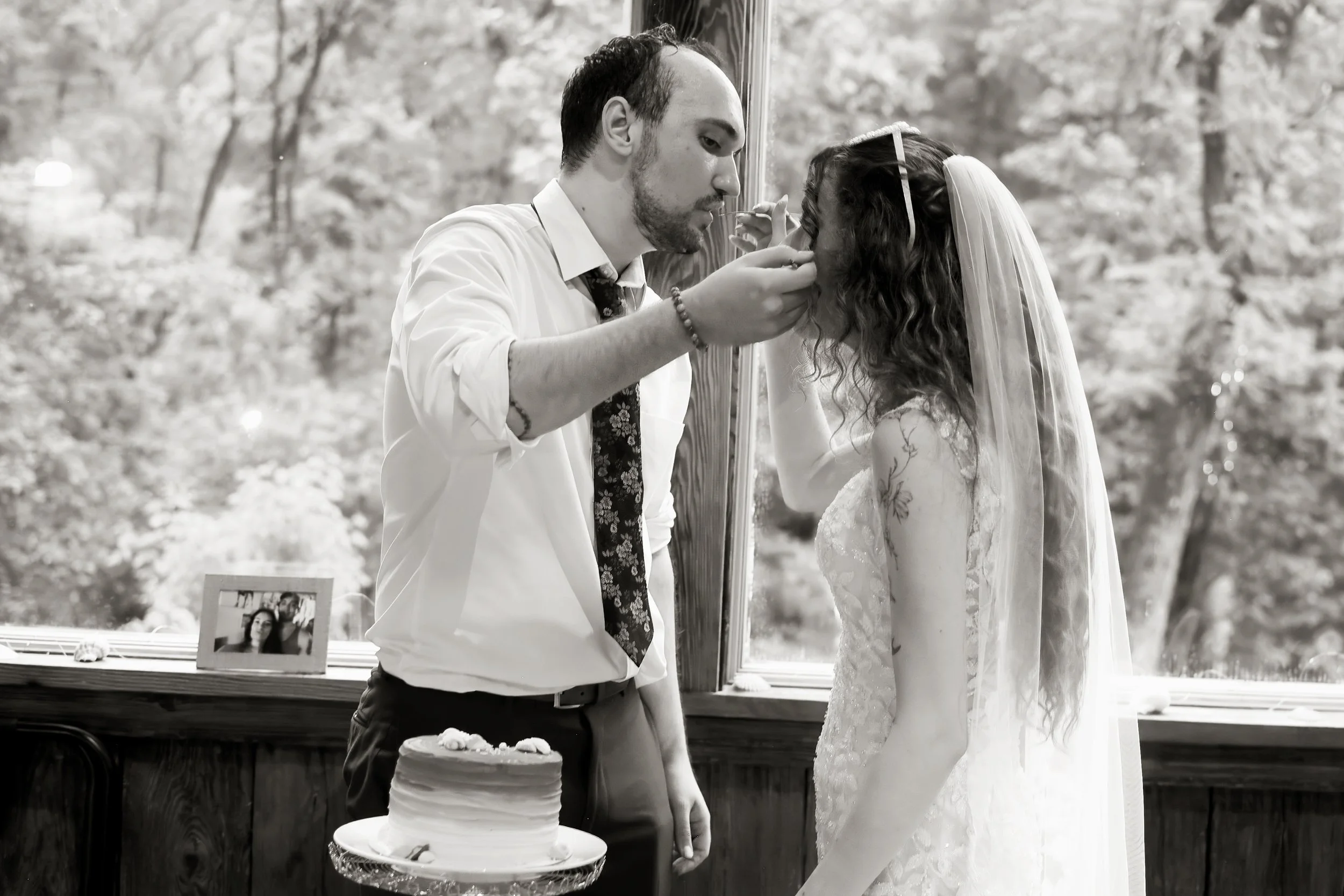 A man in a white shirt and tie is touching a woman's face. The woman is dressed in a wedding gown and veil. They are standing near a window with trees outside. A wedding cake and a framed photo are on a table in front of them.