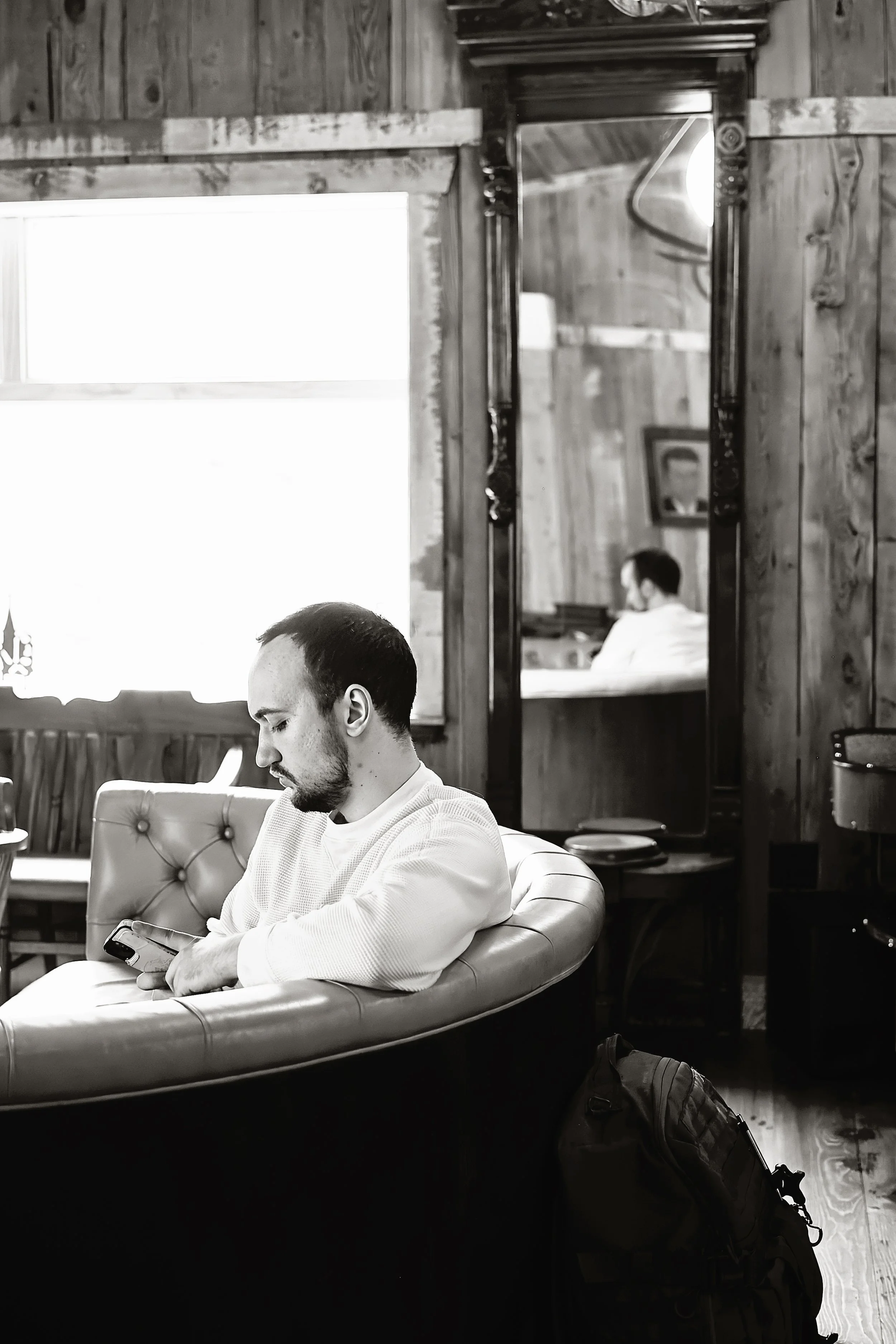 A man with short dark hair and a beard, wearing a white sweater, is sitting on a cushioned armchair in a cozy wooden interior, looking at his smartphone. A large window is behind him, with some furniture visible reflecting in a tall mirror on the wal