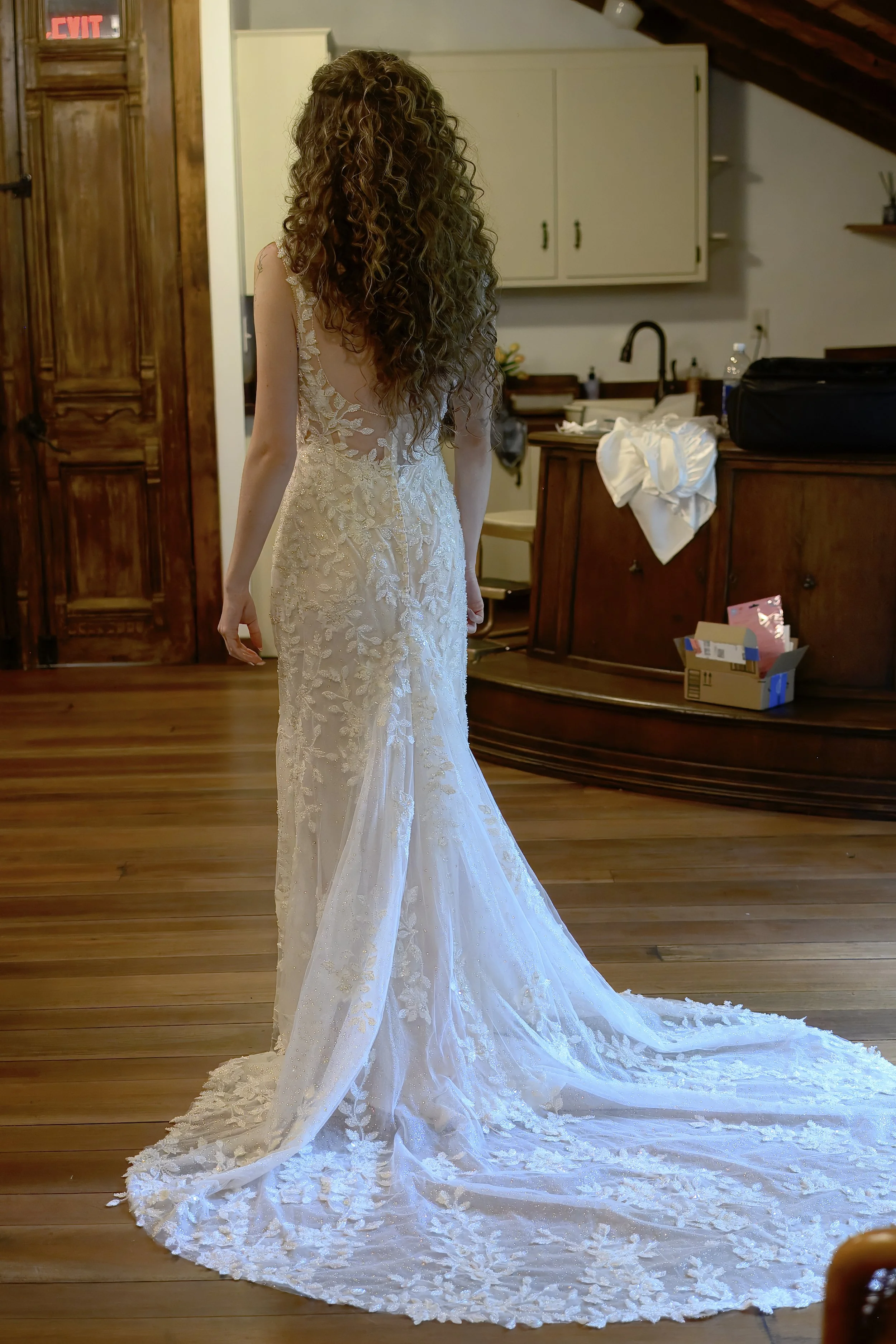 A woman wearing an elegant white wedding gown with lace and embroidery detailing, standing on a wooden floor in a cozy room with wooden furniture and a white cabinet. Her face is not visible as she looks away. The gown has a long train and floral pat