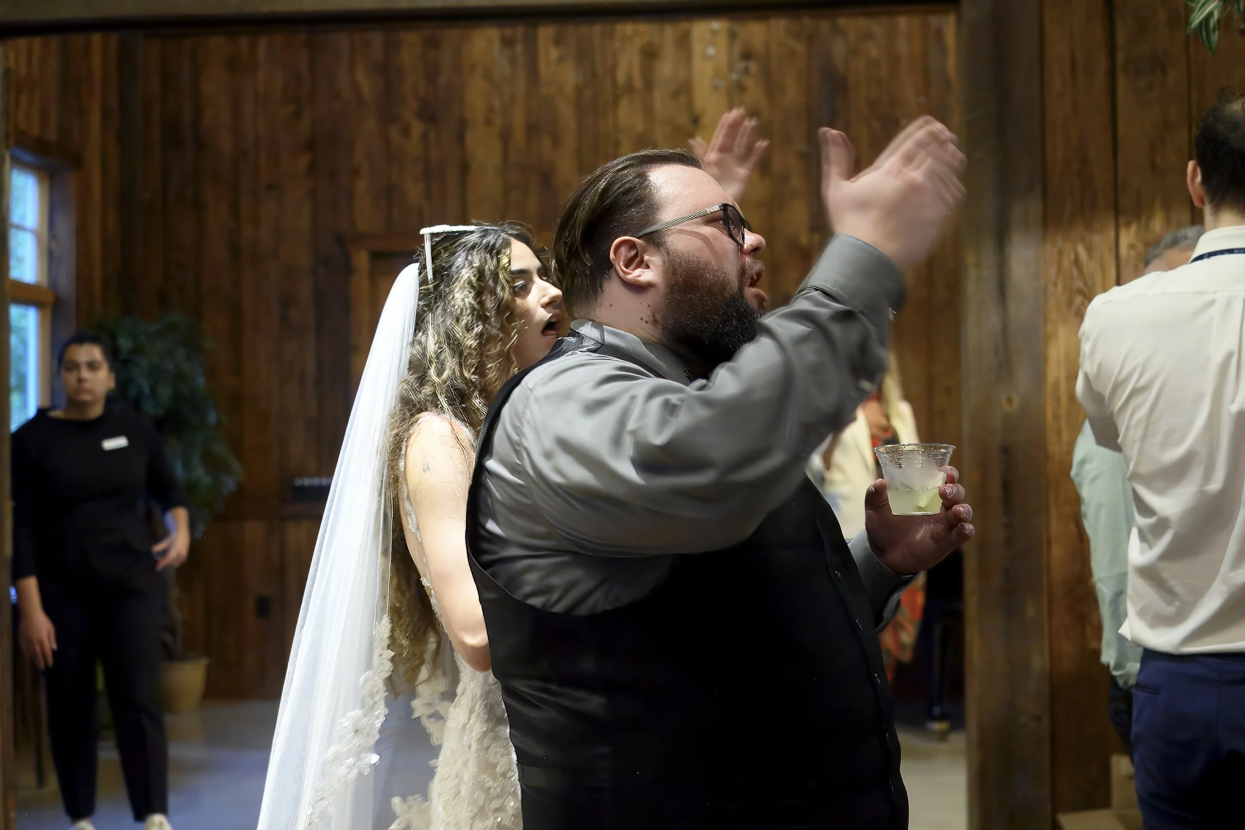 Man with glasses raising his hand and holding a drink during a wedding celebration, with a bride standing behind him, in a wooden interior.