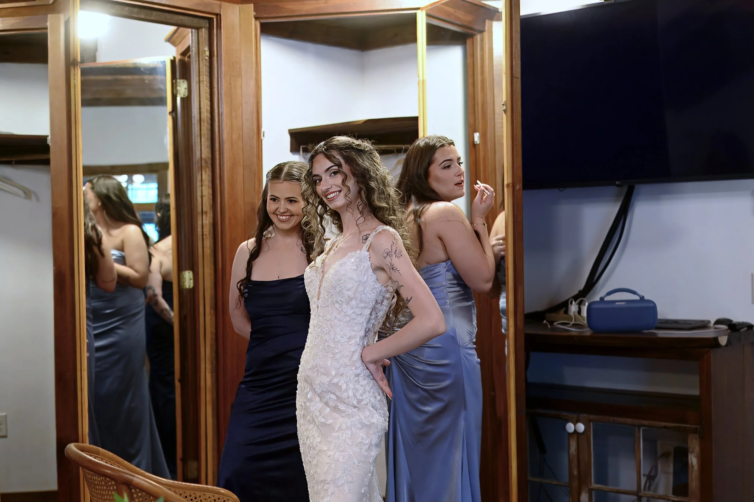 Three women in formal dresses standing in front of a mirror, smiling, with a wooden frame, in a room with a dark wall and a TV.