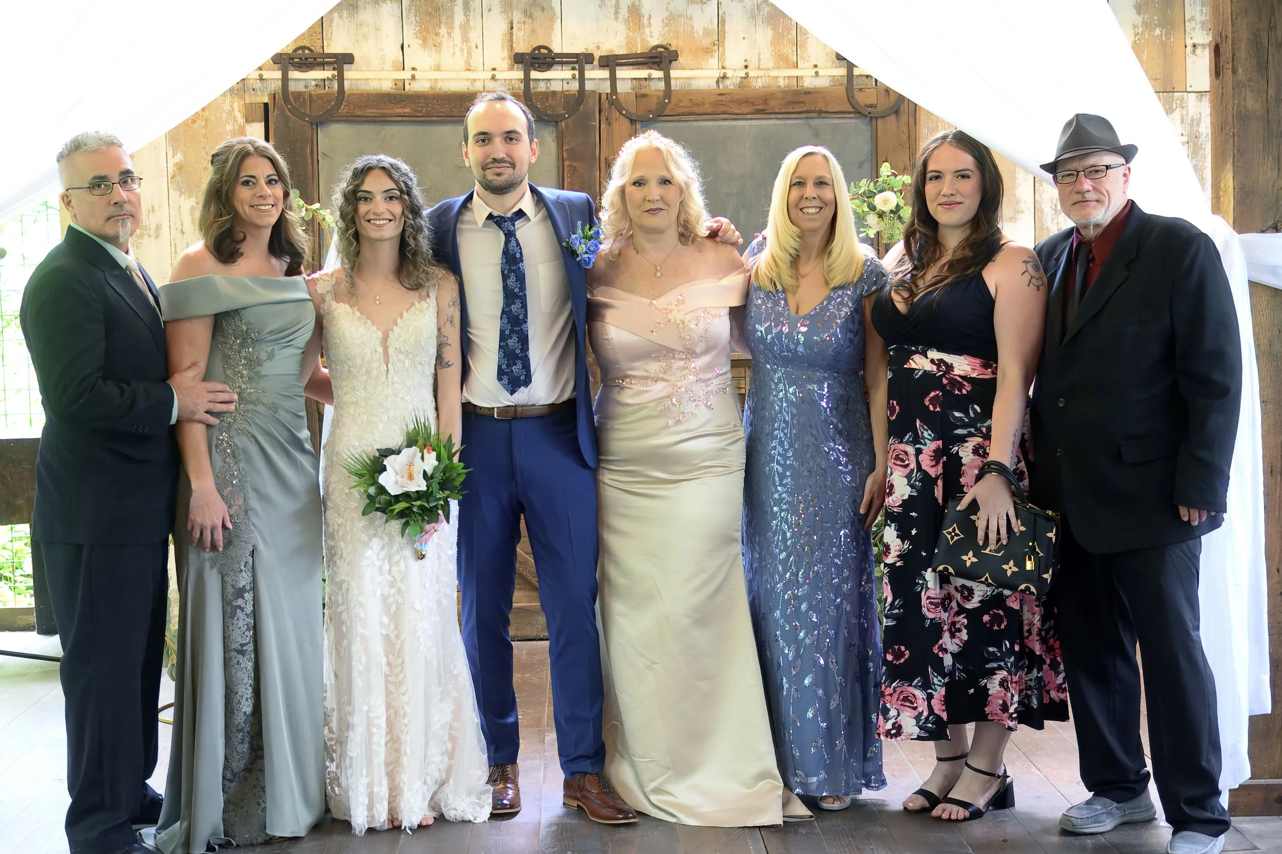 Group of nine people standing together at a wedding, posing for a photo inside a rustic venue with wooden walls and greenery visible in the background.