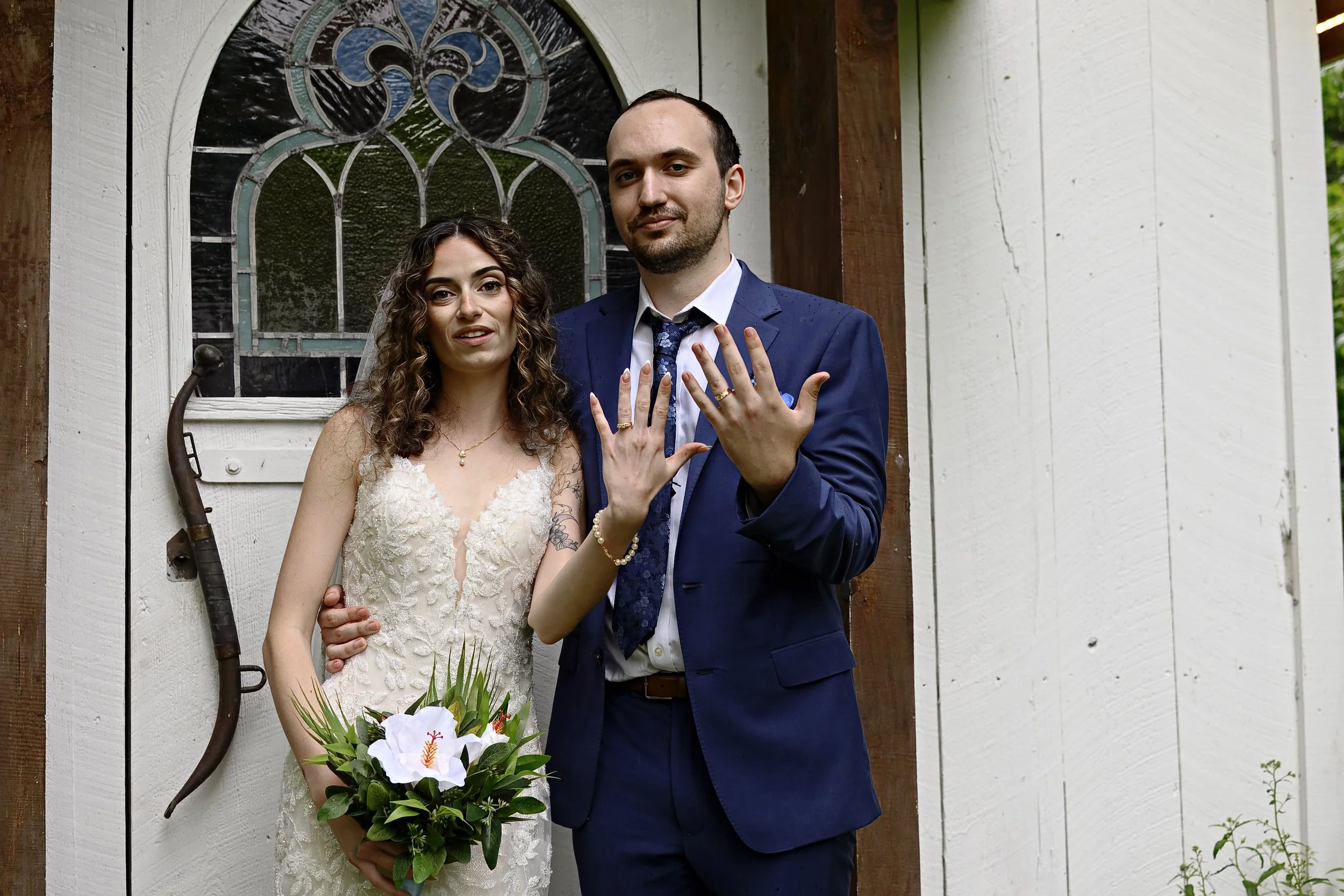 A newly married couple standing outside a white wooden building with a stained glass window, holding hands and showing their wedding rings. The bride is holding a bouquet of white and orange flowers, and the groom is dressed in a blue suit and tie.