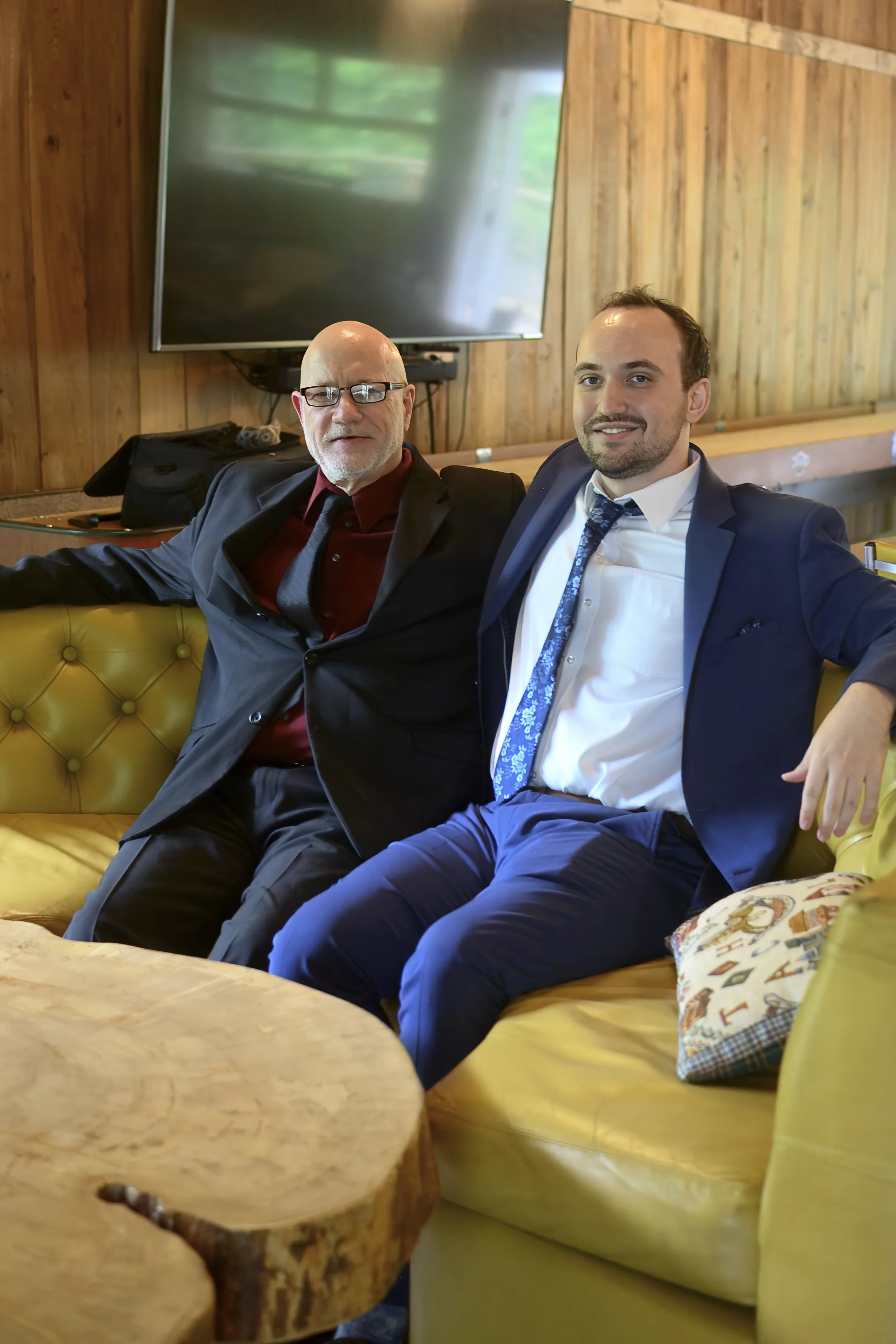 Two men sitting on a yellow leather sofa in a wooden-paneled room, smiling at the camera. One is bald with glasses, wearing a black suit and red shirt, and the other has short dark hair, wearing a blue suit with a patterned tie. A large flat-screen T