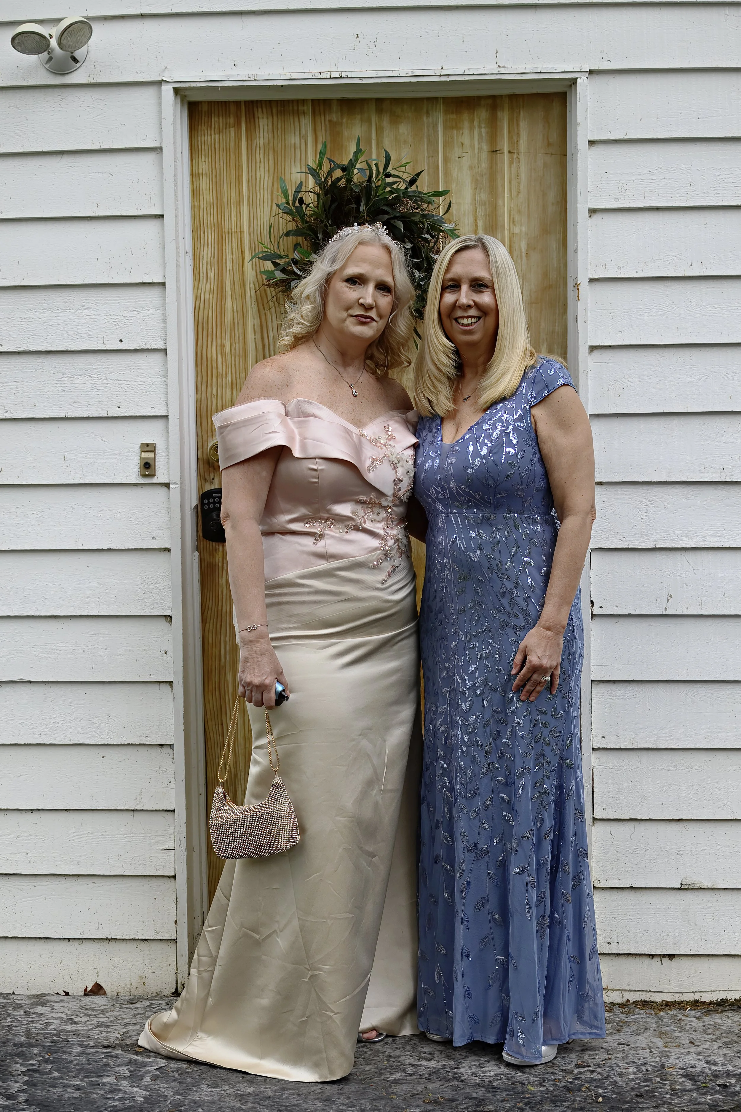 Two women standing close together in front of a wooden doorway, posing for a photo. One woman is wearing a pink off-shoulder gown with a tiara, and the other is wearing a blue dress with a leaf pattern.
