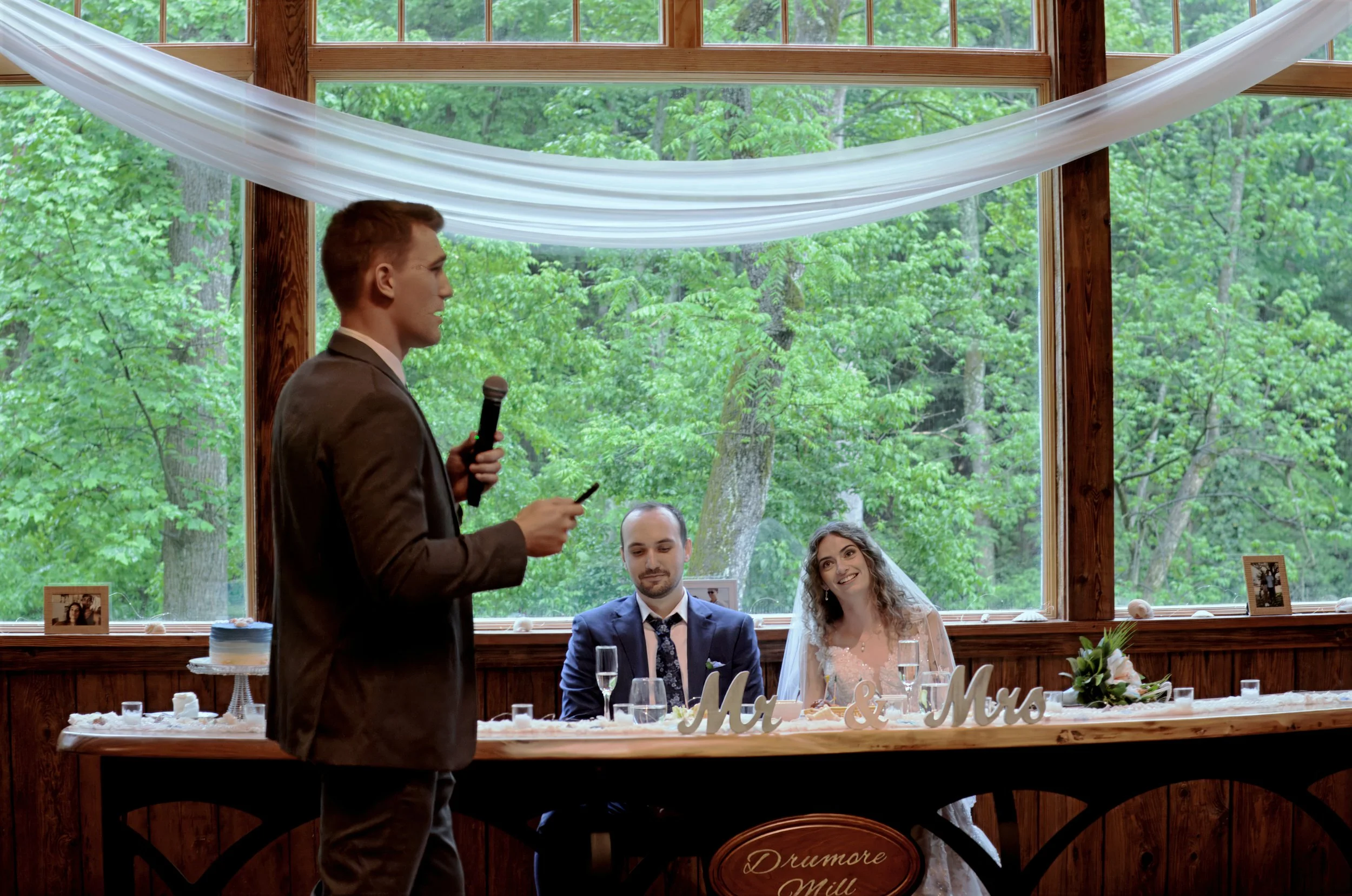 A man in a brown suit is giving a speech at a wedding reception, holding a microphone and a phone, while three guests in formal attire—two men and a woman—are sitting at a table decorated with a sign that says 'Mr & Mrs' and various glasses and decor
