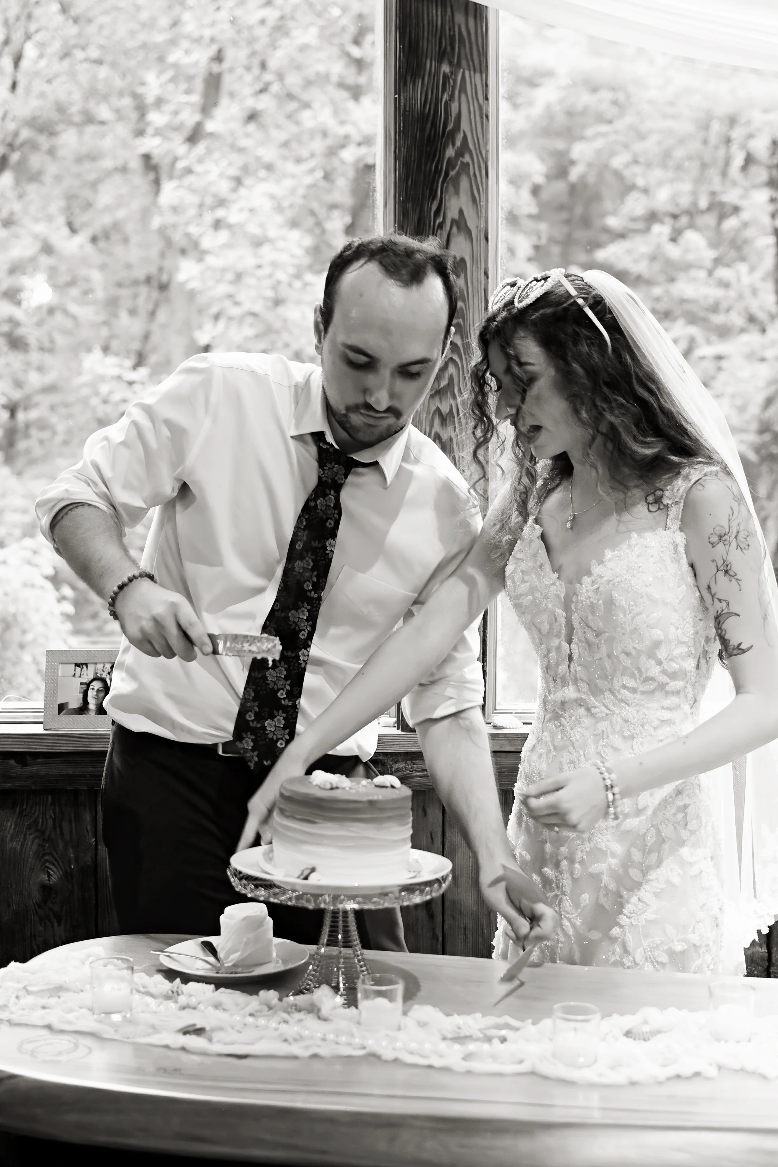 A bride and groom cutting a wedding cake together during their wedding reception, with a table of desserts and a framed photograph in the background.