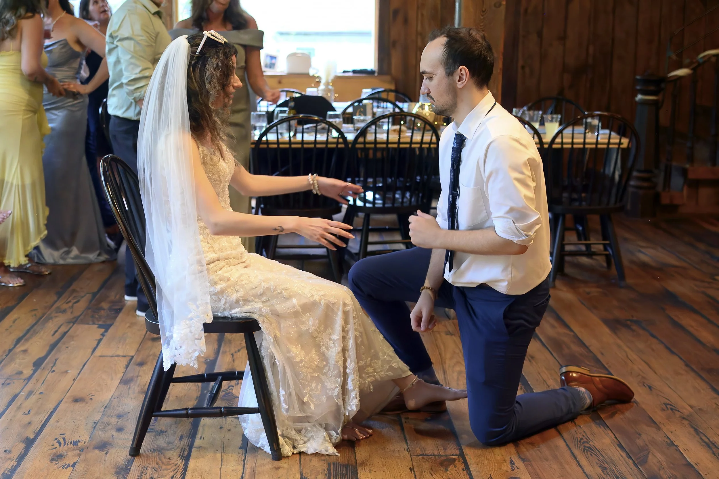 A wedding ceremony in a rustic wooden venue, with a bride seated and a groom kneeling in front of her, holding her hand. The bride wears a lace wedding dress and veil, while the groom wears a white shirt with rolled-up sleeves and a dark tie. Guests 