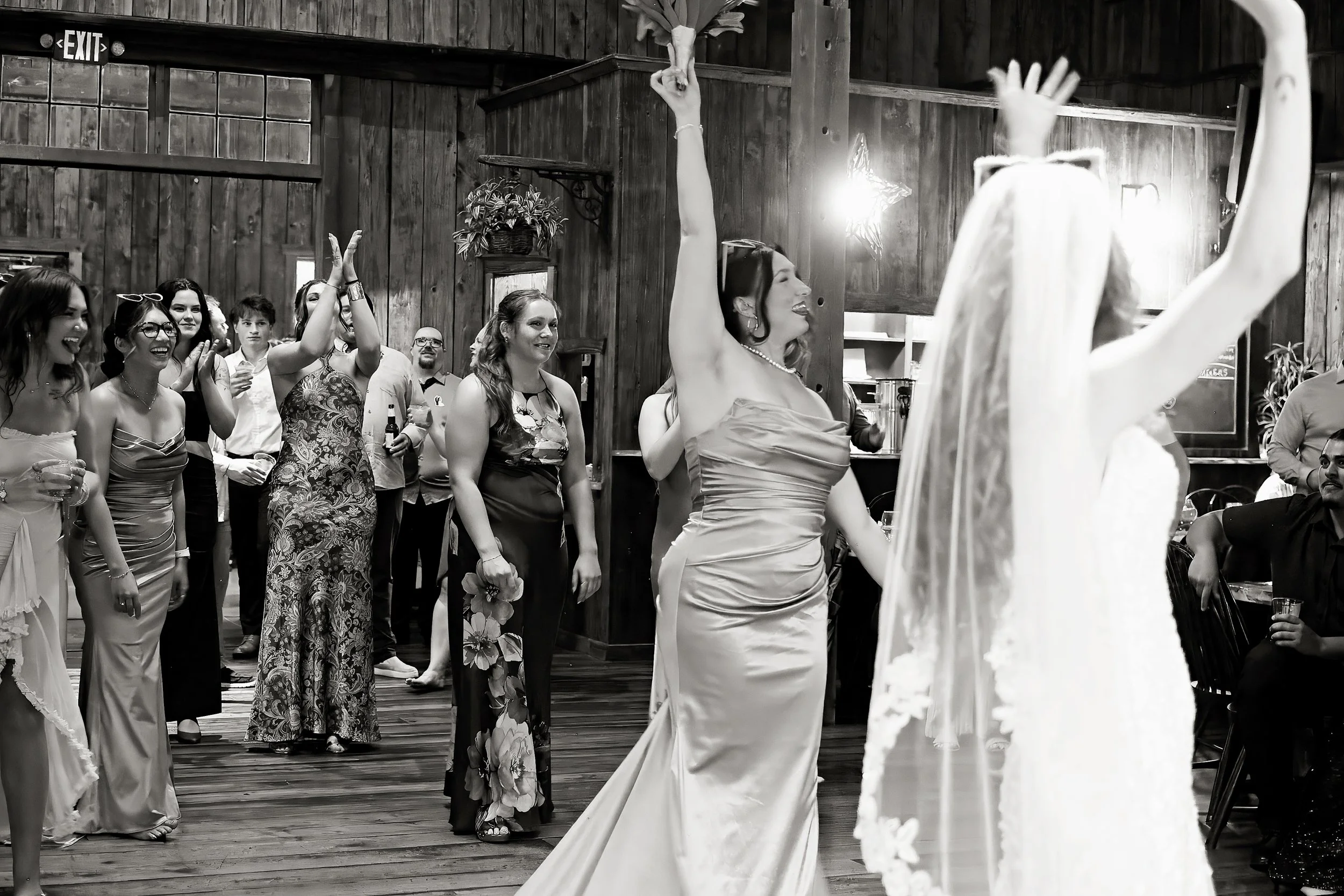 A group of women in formal dresses dancing and smiling at an indoor event with wooden walls.
