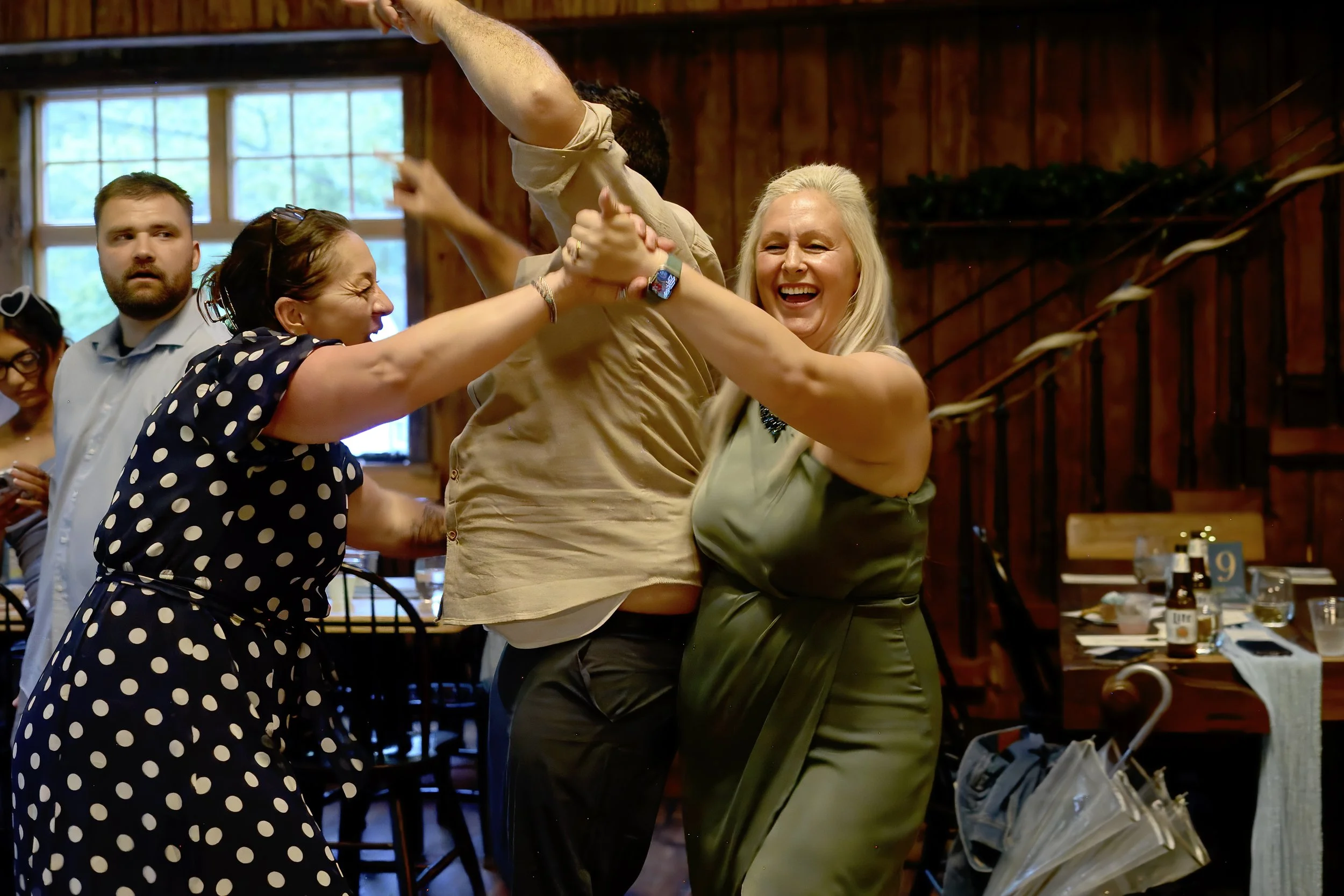 Two women and a man dancing together in a rustic wooden room, smiling and holding hands, while other people look on in the background.