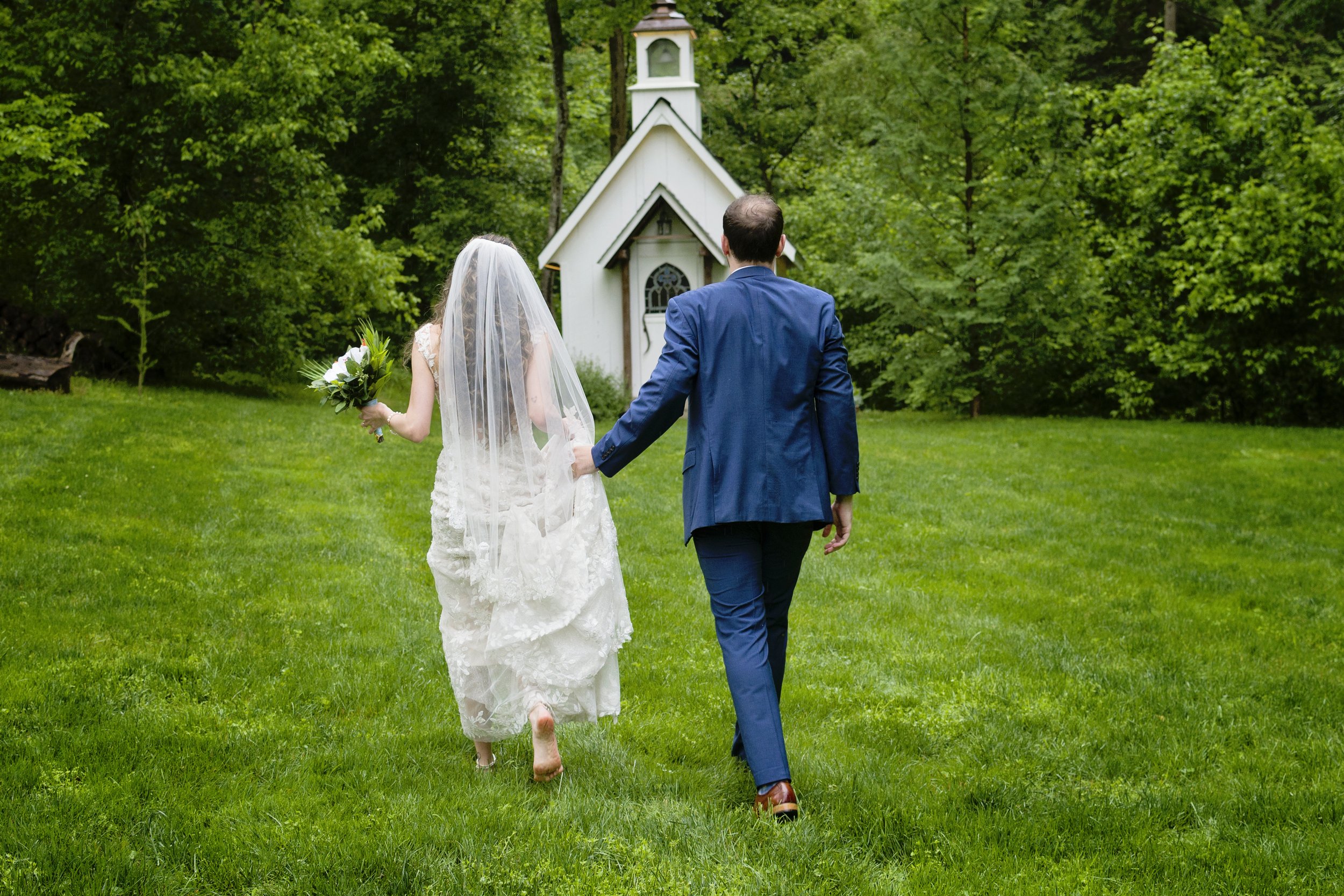 A bride and groom walking hand in hand towards a small white chapel in a lush green outdoor setting.