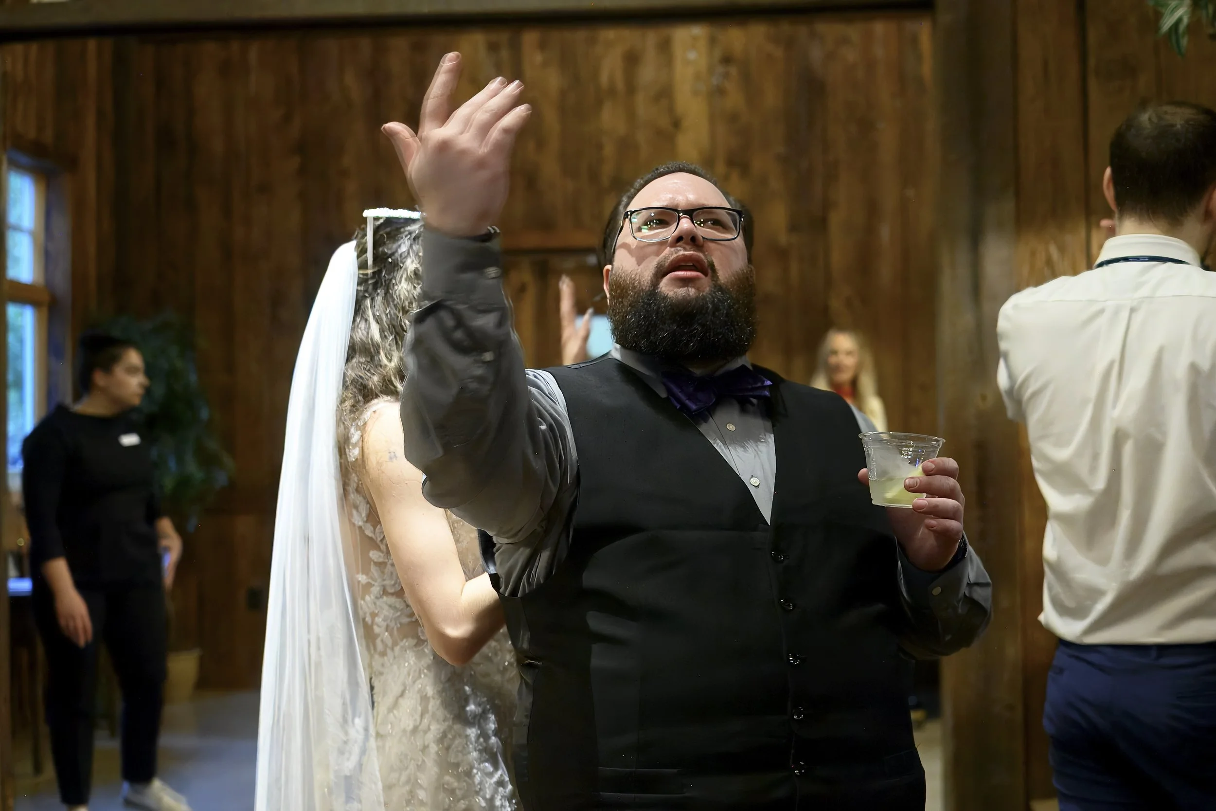 A man with glasses and a beard, dressed in a tuxedo, holding a drink, standing close to a woman in a wedding dress at a celebration or wedding reception.