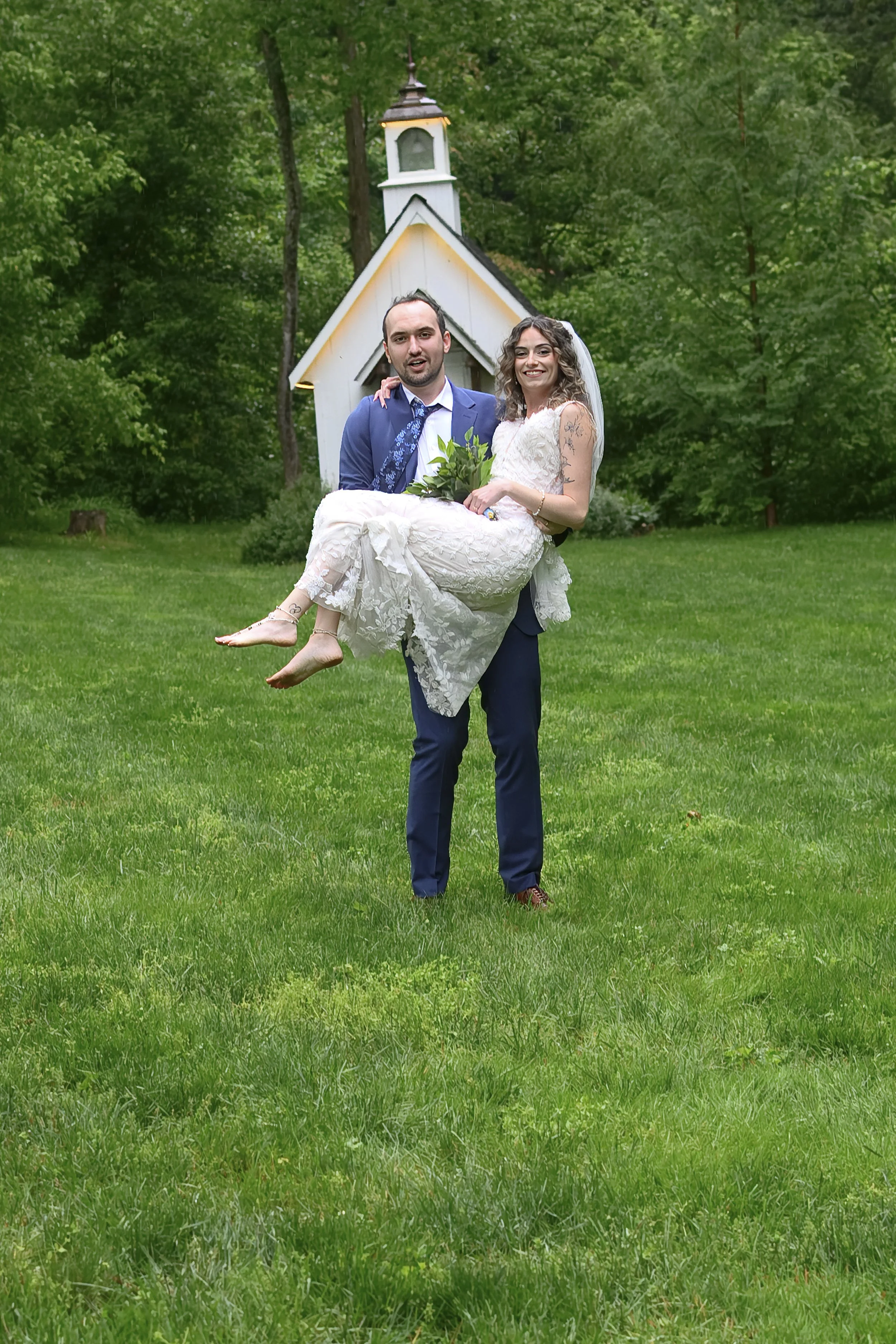 A man and woman in wedding attire standing on a grassy lawn in front of a small white chapel surrounded by trees. The man is holding the woman in his arms, and they are smiling at the camera.