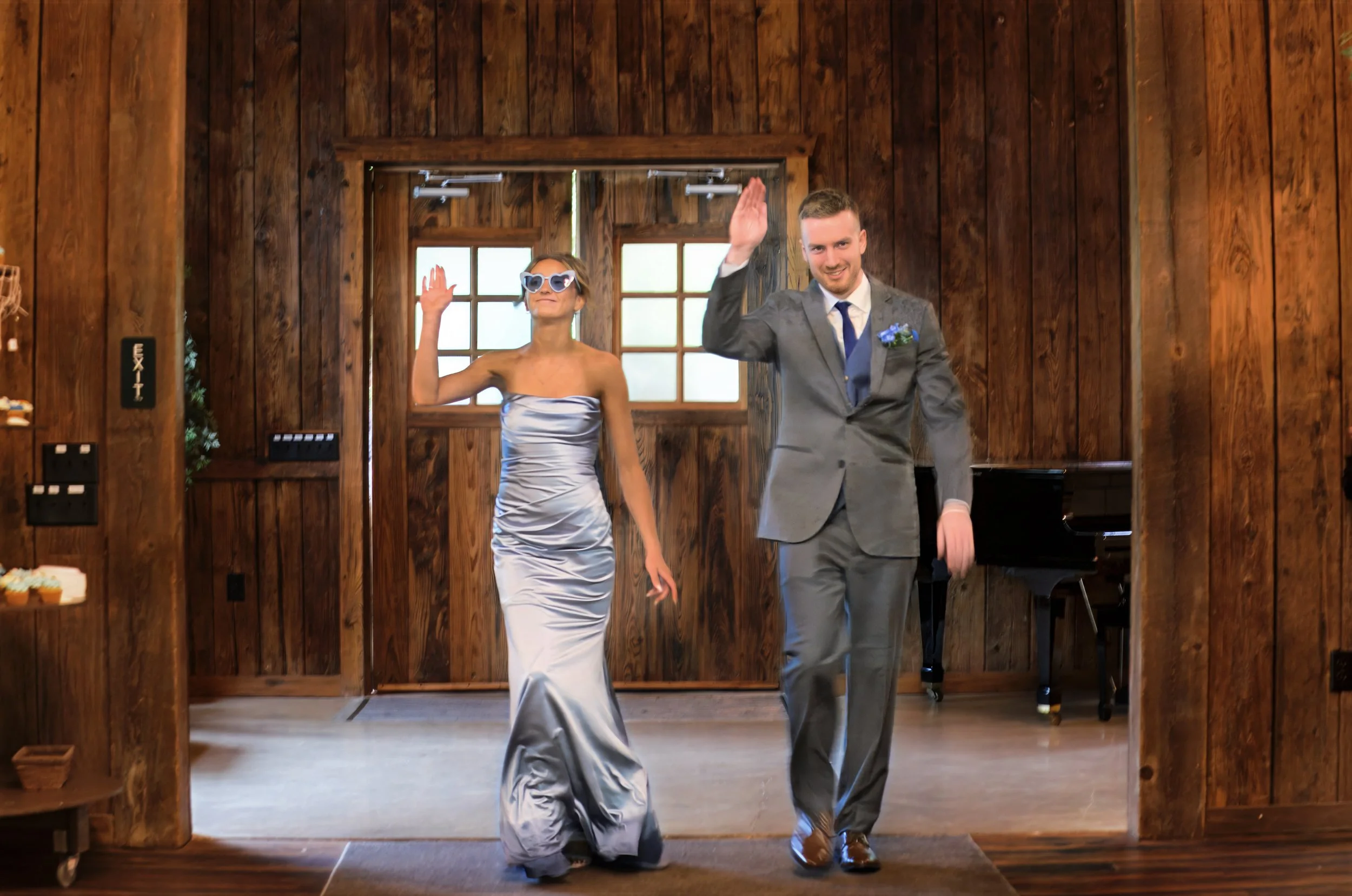 A woman in a silver satin strapless gown and sunglasses and a man in a gray suit with a blue tie and boutonniere walk down a wooden stage while waving to the audience.