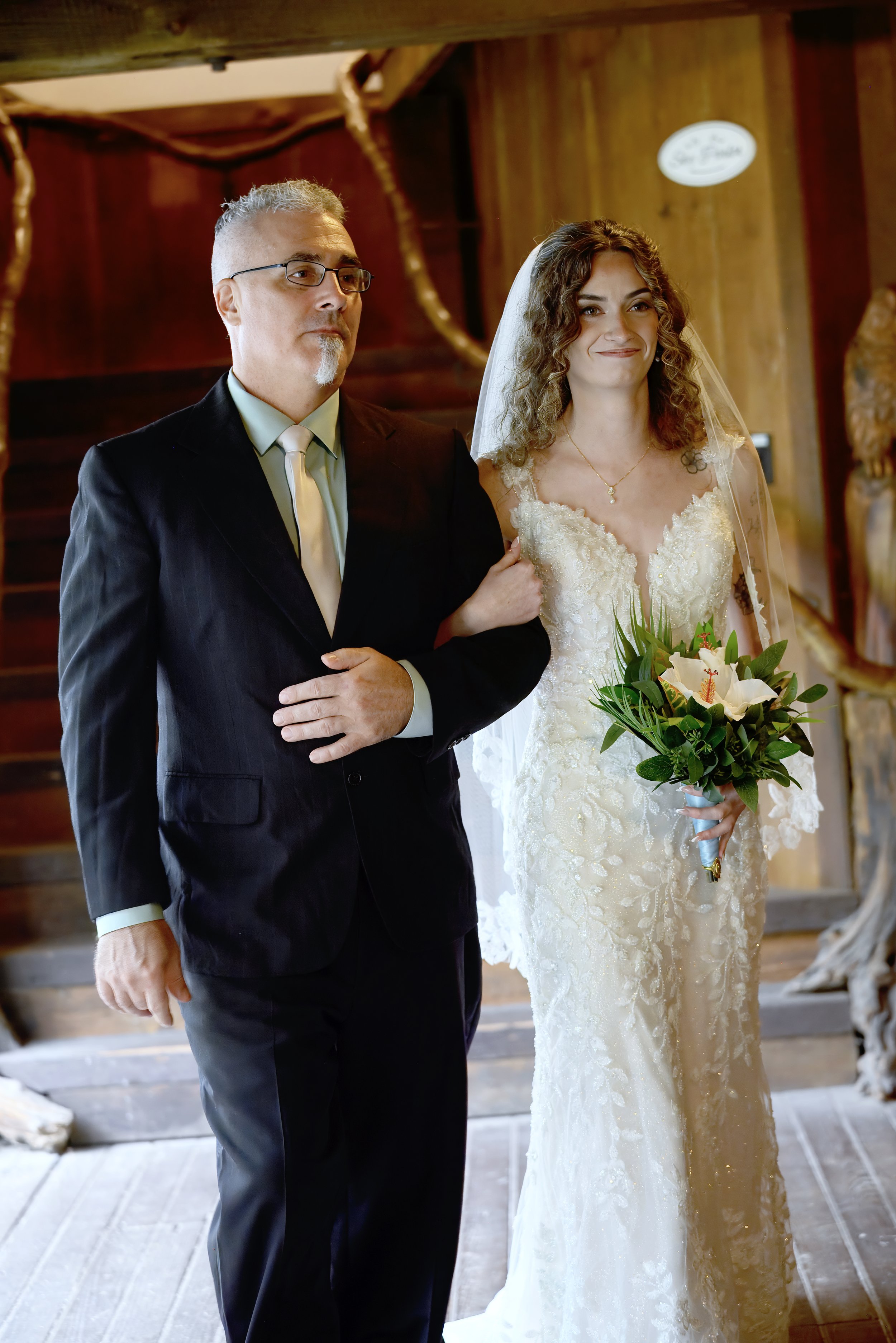 A bride with curly brown hair wearing a white lace wedding dress and holding a bouquet of white lilies and greenery, standing beside a man in a black suit with glasses and gray hair, inside a wooden venue with warm lighting.