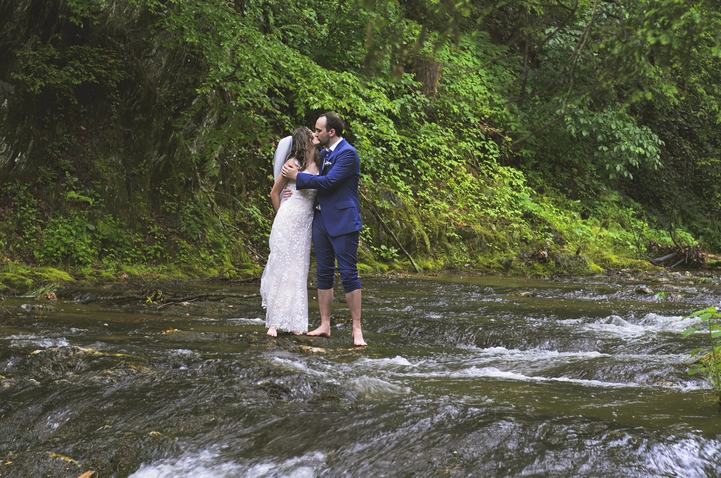 A newlywed couple stands barefoot in a shallow creek, sharing a kiss surrounded by lush green foliage.