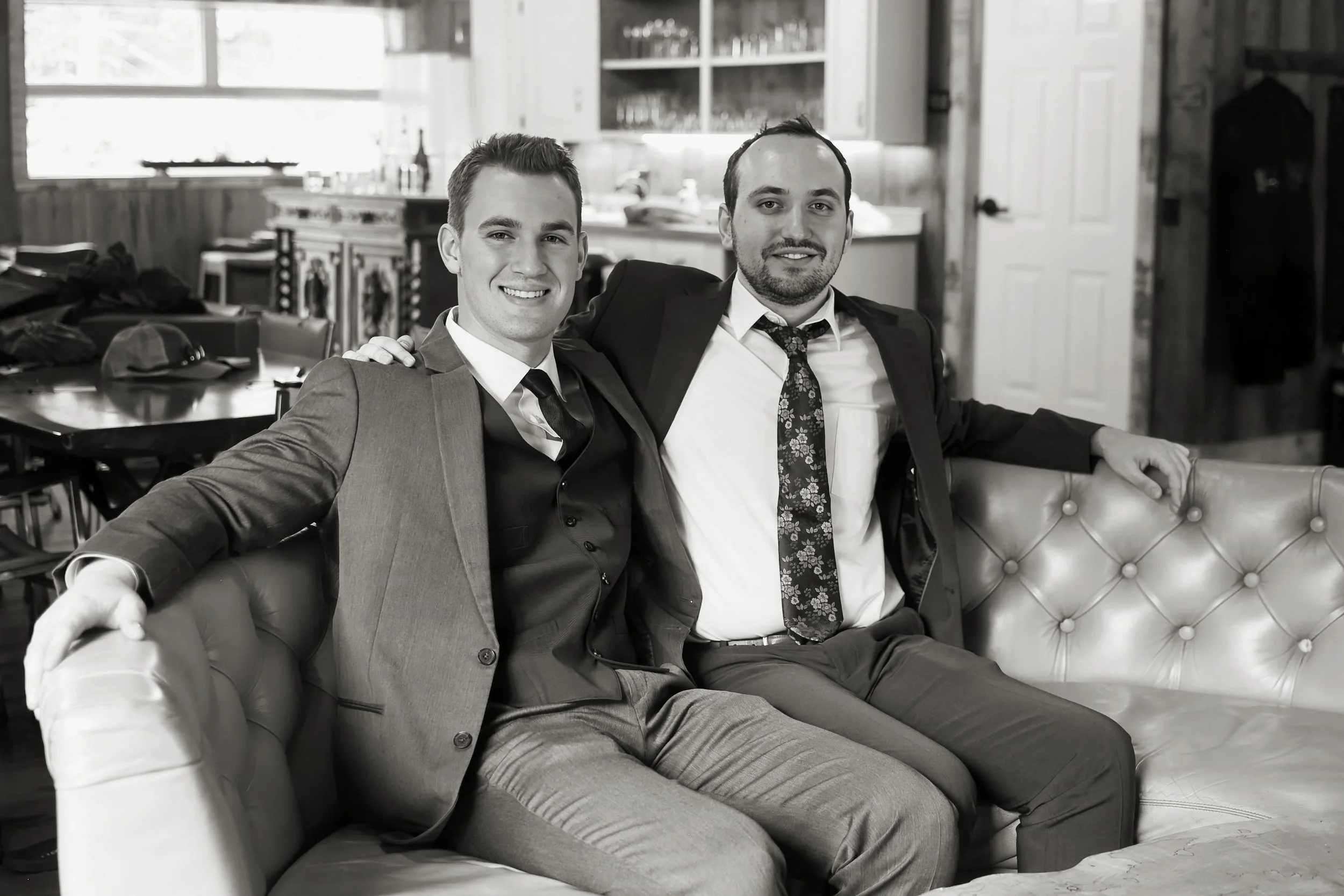 Two men in suits sitting on a tufted leather couch in a kitchen, smiling at the camera. One man has his arm around the other.